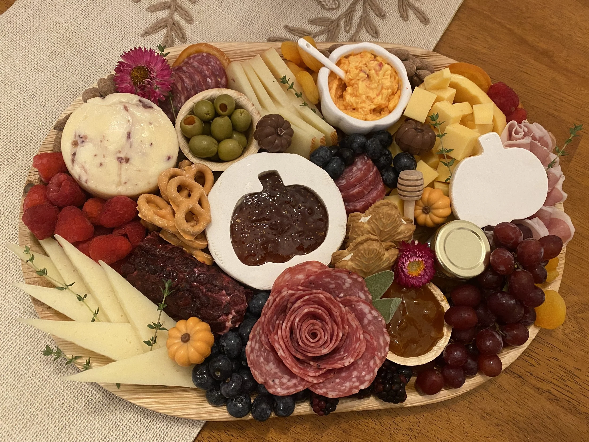 A round wooden cheese board with assorted cheeses, grapes, berries, cured meats, jams, crackers, and decorative flowers.