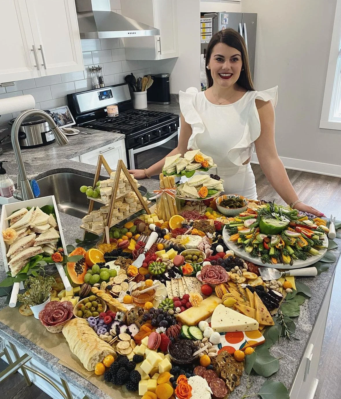 A woman in a white dress stands behind a kitchen island decorated with a variety of food items including sandwiches, cheeses, fruits, vegetables, and charcuterie, smiling at the camera.