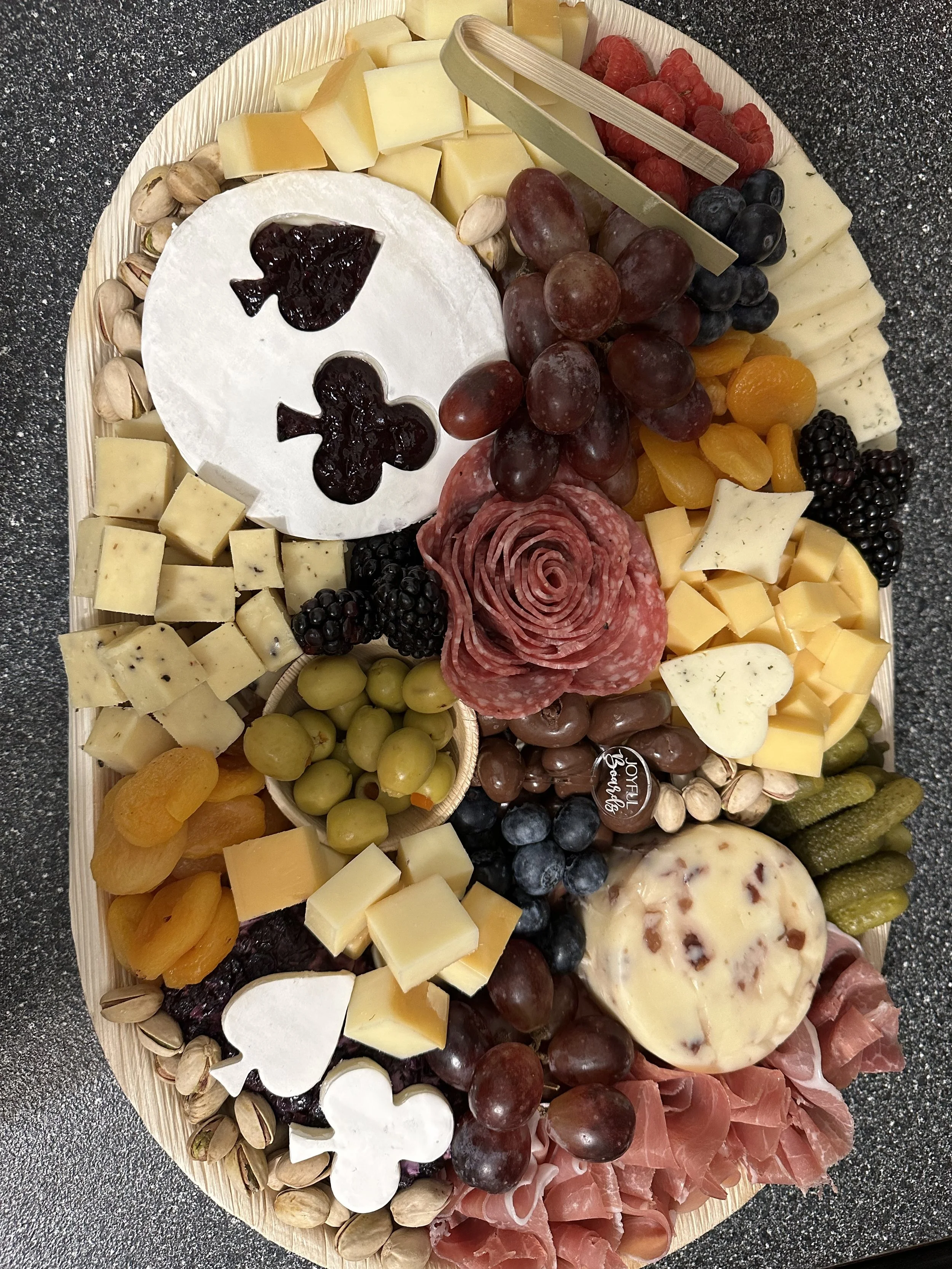A cheese and fruit platter with a carved cheese block in the shape of a ghost, assorted cheeses, grapes, berries, dried apricots, pistach nuts, and slices of meats, arranged on a wooden tray on a dark countertop.