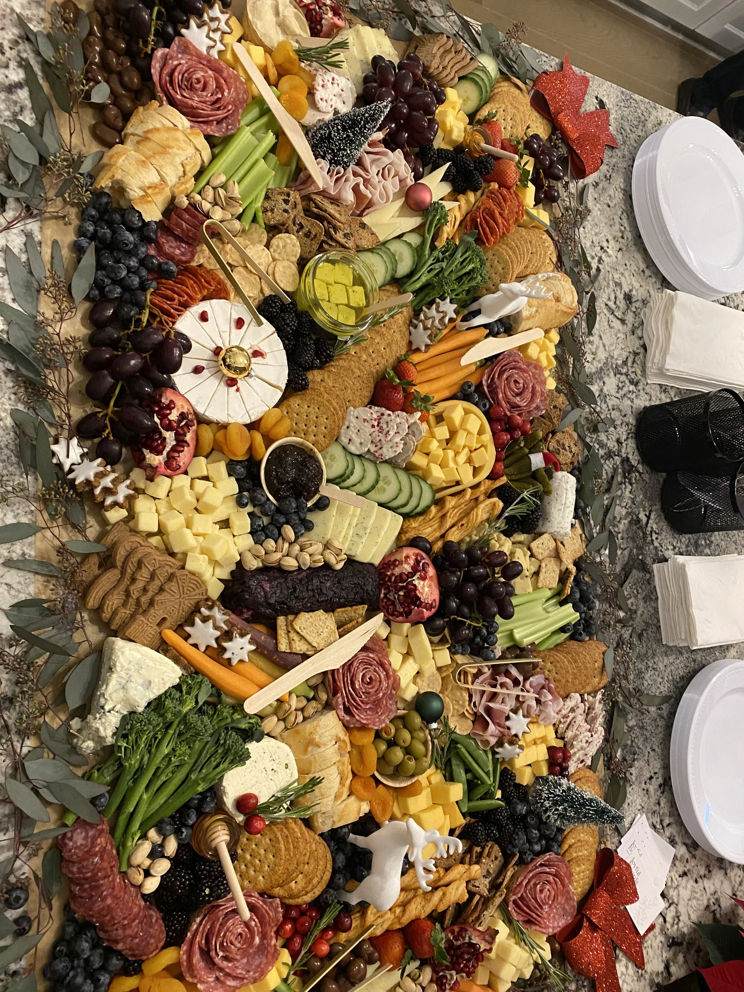 A large charcuterie board featuring assorted cheeses, meats, crackers, grapes, berries, vegetables, and decorative holiday ornaments on a granite countertop.