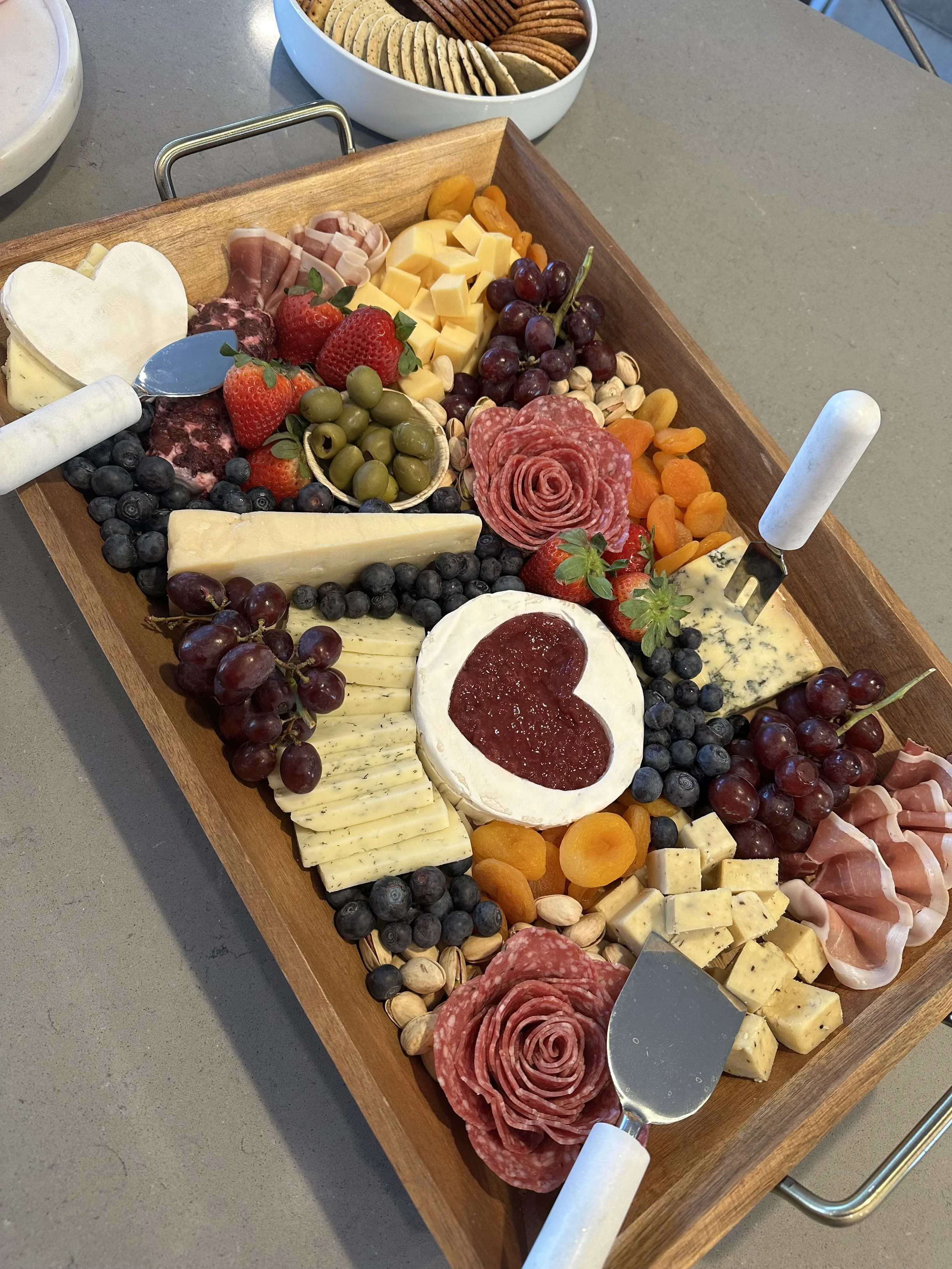 A wooden charcuterie board with various cheeses, meats, grapes, strawberries, and almonds. There are also cheese knives present.