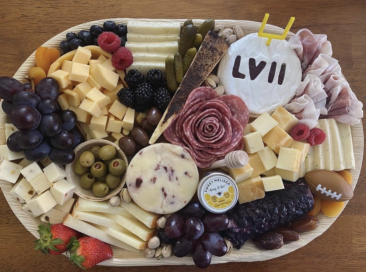 A football-themed cheese and fruit platter with grapes, cheese cubes, raspberries, blackberries, green olives, pickles, sliced meats, a wheel of cheese labeled "Sweet Melissa Honey & Blueberry," a round cheese with a floral pattern, a chocolate-cover