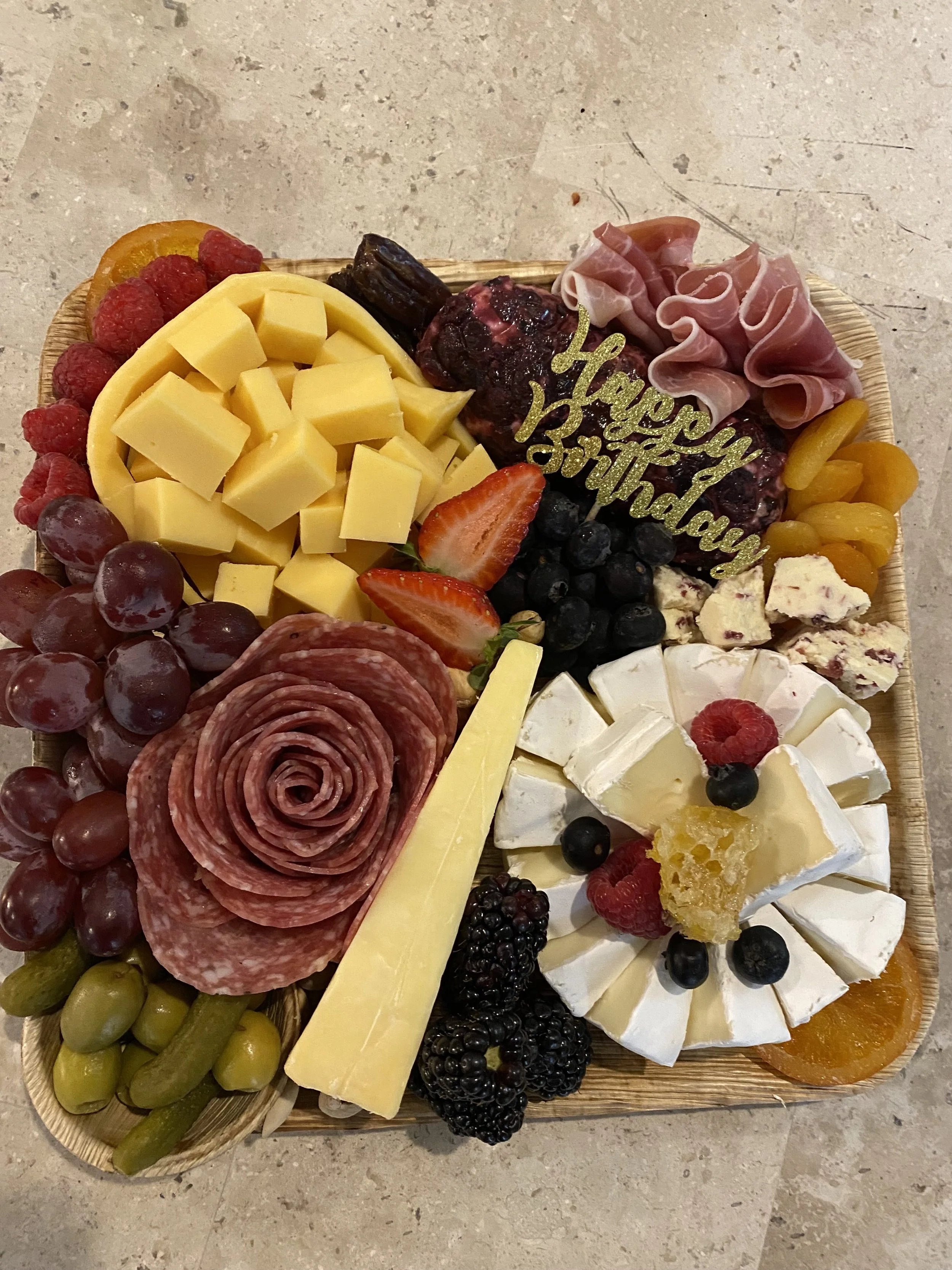 A cheese and charcuterie platter with various cheeses, meats, fresh and dried fruits, and a "Happy Birthday" decoration.