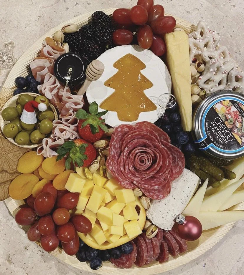 A cheese and charcuterie board with grapes, strawberries, blueberries, blackberries, slices of cheese, salami, cheese dip, greens, and holiday decorations like a small Santa hat and pink bauble.