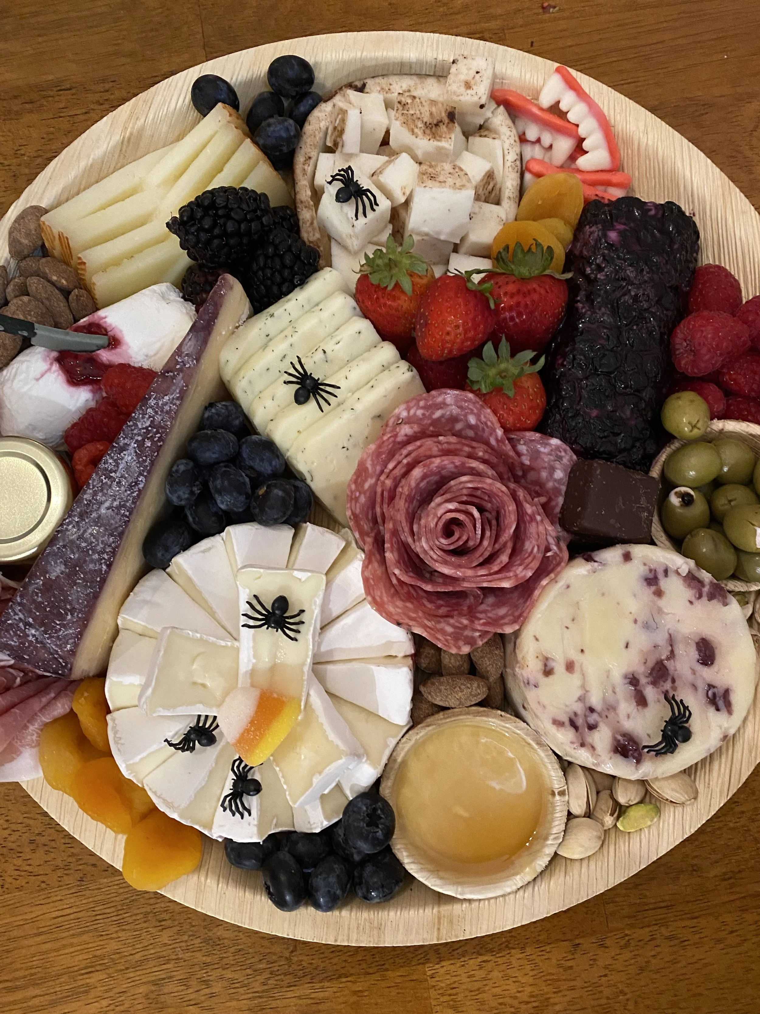 A cheese platter with various cheeses, grapes, strawberries, crackers, cured meats, berries, and garnishes on a round wooden board.
