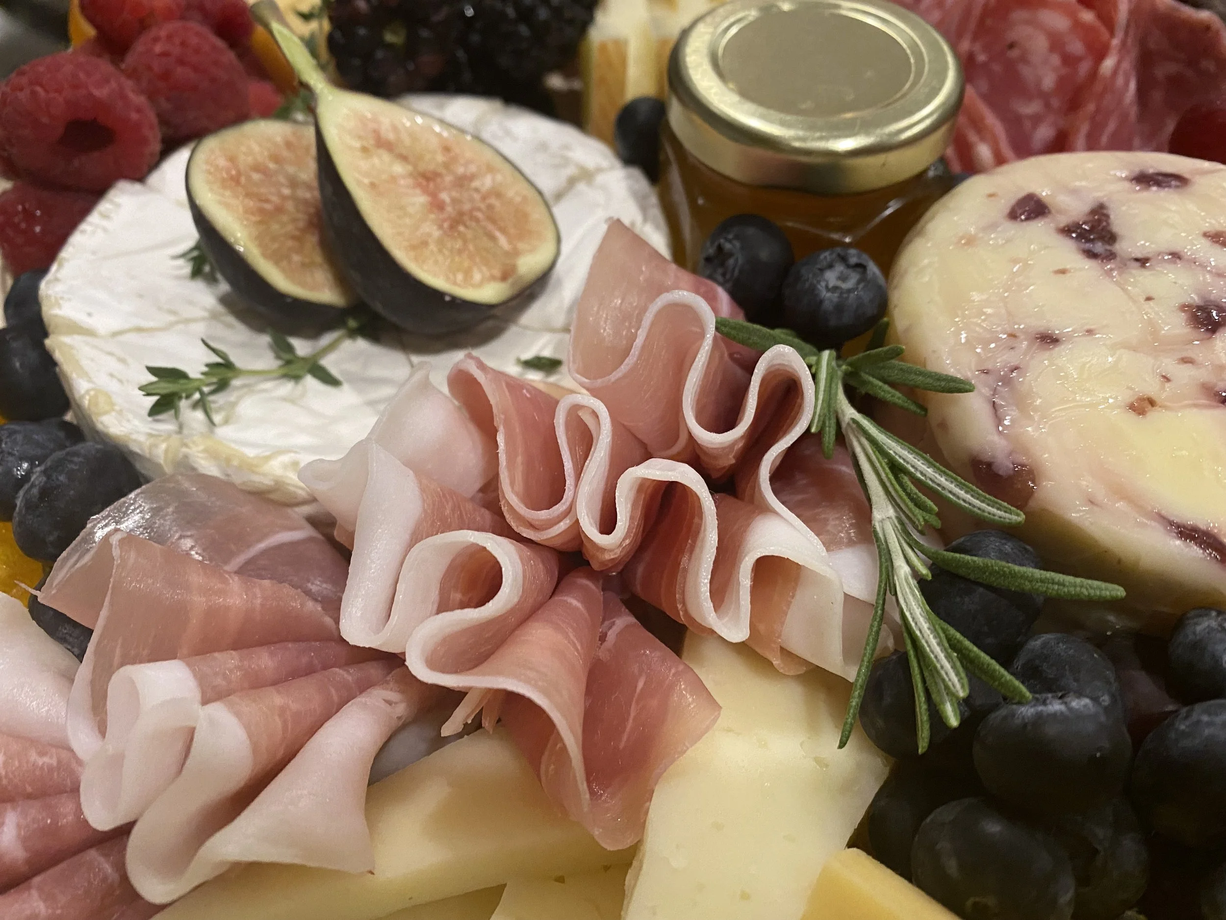 Close-up of a cheese and charcuterie platter with fresh figs, blueberries, honey jar, cured meats, and rosemary.