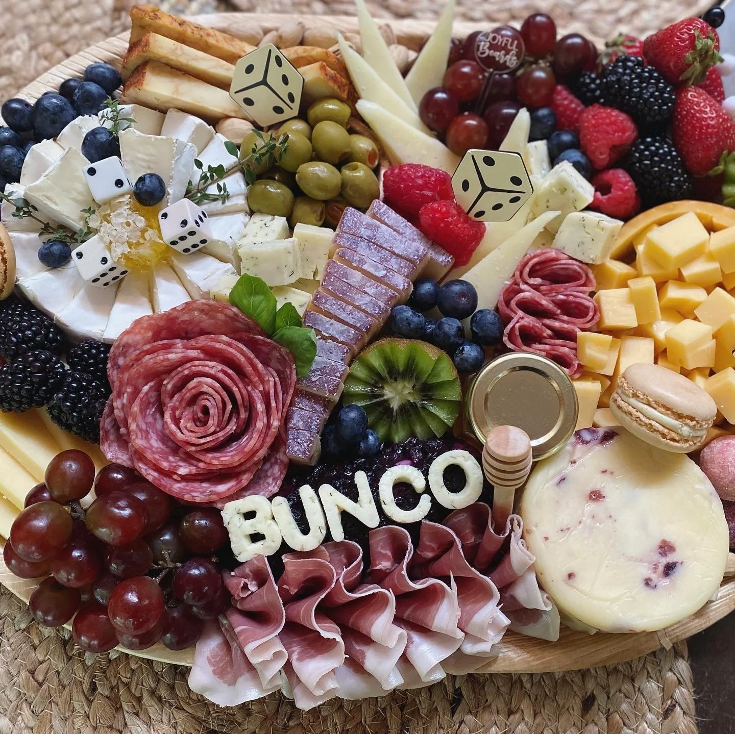 Cheese, grapes, strawberries, raspberries, blackberries, melon slices, salami, prosciutto, cheese cubes, macarons, bowl with gold lid, dice, small jar, and decorative elements on a wooden platter.