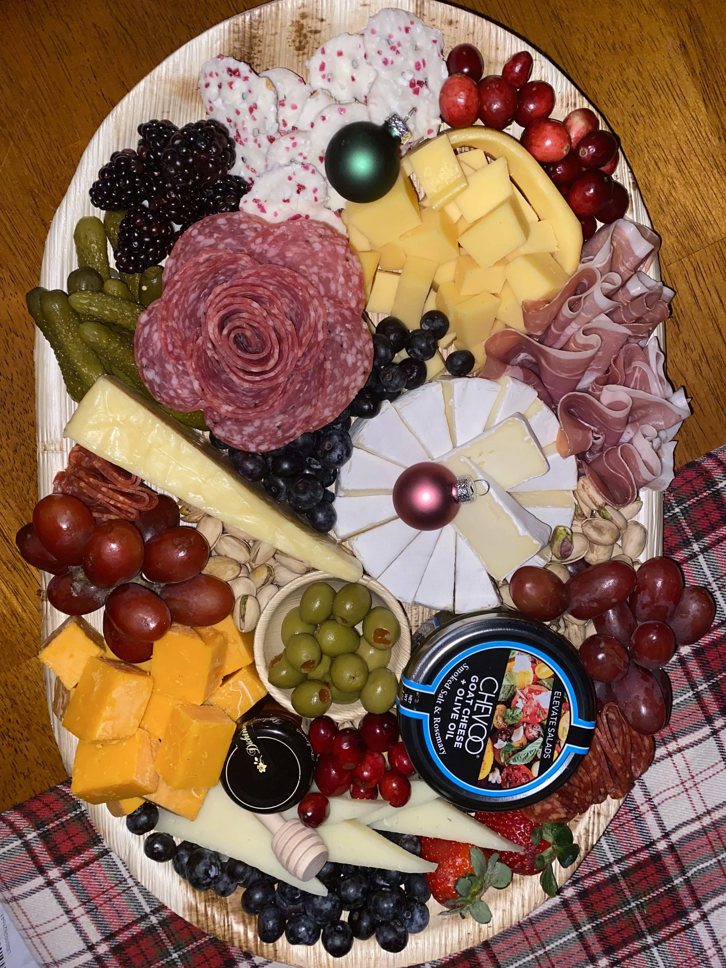 A wooden platter with assorted cheeses, grapes, strawberries, blueberries, blackberries, melon slices, cured meats, crackers, and holiday ornaments, on a plaid cloth.