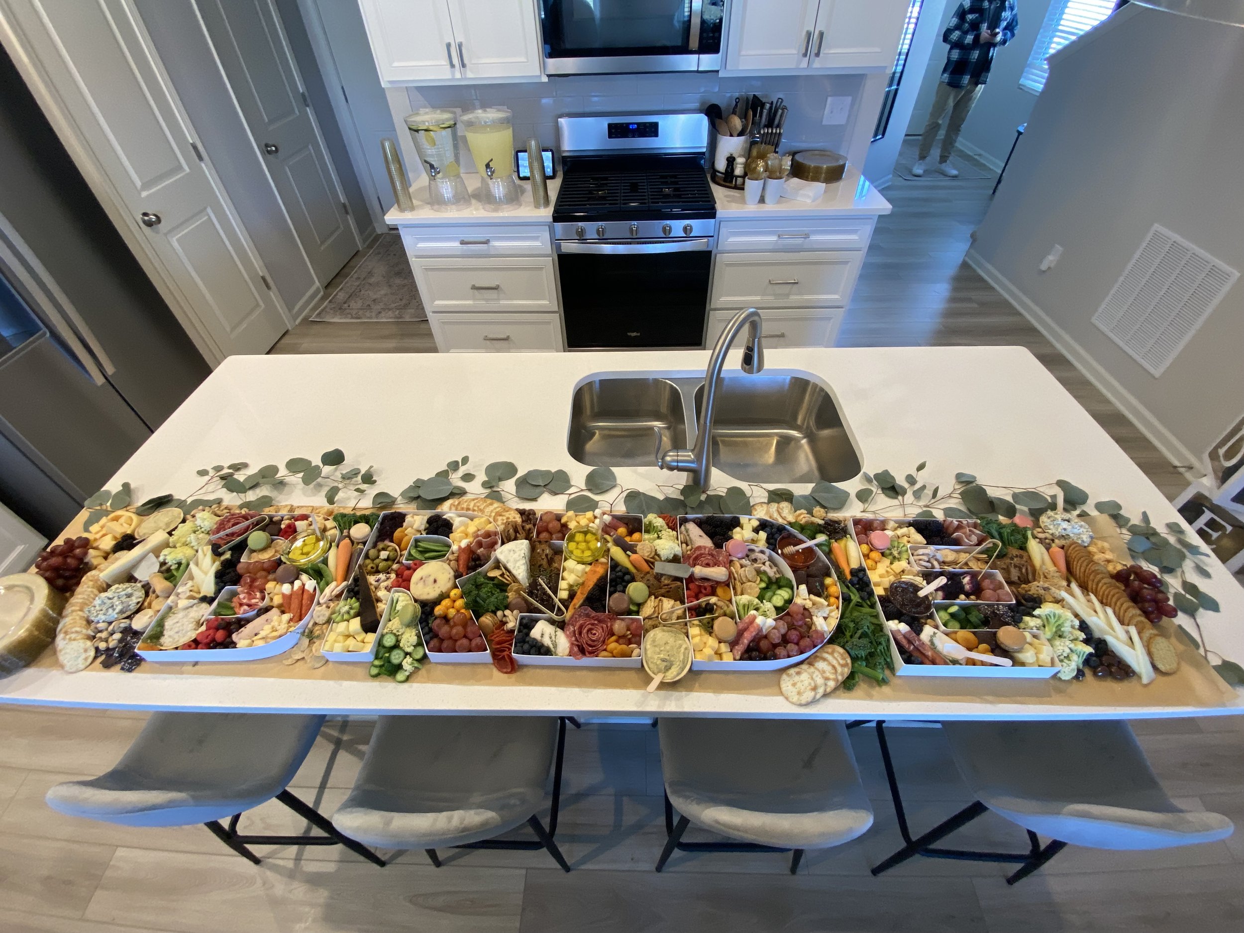 Overhead view of a large charcuterie board filled with various cheeses, cured meats, fruits, vegetables, crackers, and small bowls of condiments, set on a kitchen table. In the background, a kitchen with white cabinets, a stove, and a stone counterto