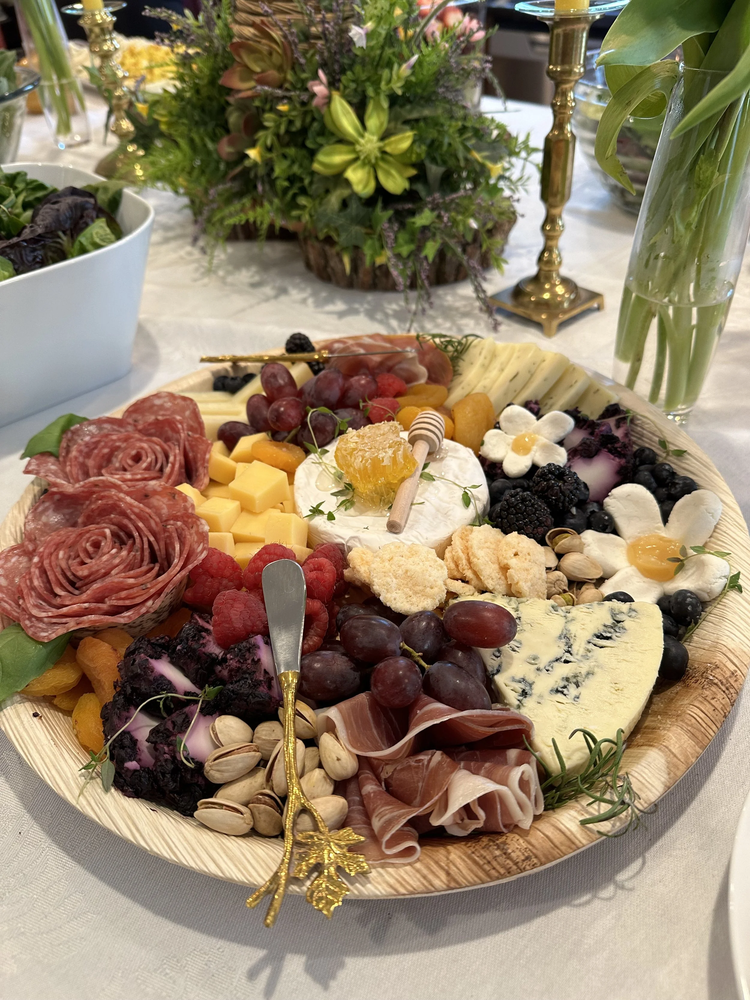 A cheese and charcuterie platter with assorted cheeses, grapes, berries, meats, and nuts, decorated with a gold pine cone ornament. In the background are floral arrangements and candlesticks.