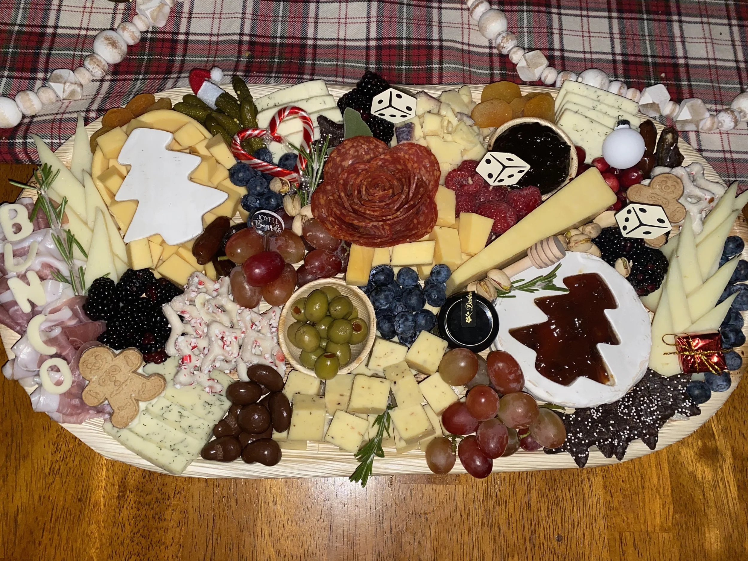 A large platter of assorted cheeses, meats, grapes, berries, and holiday decorations on a wooden table with a red plaid tablecloth in the background.