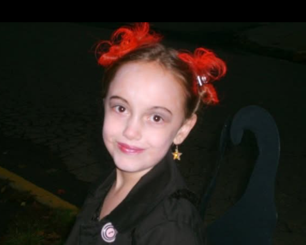 A young girl with red, curly hair styled in buns, wearing a black shirt and star-shaped earrings, smiling outdoors at night.