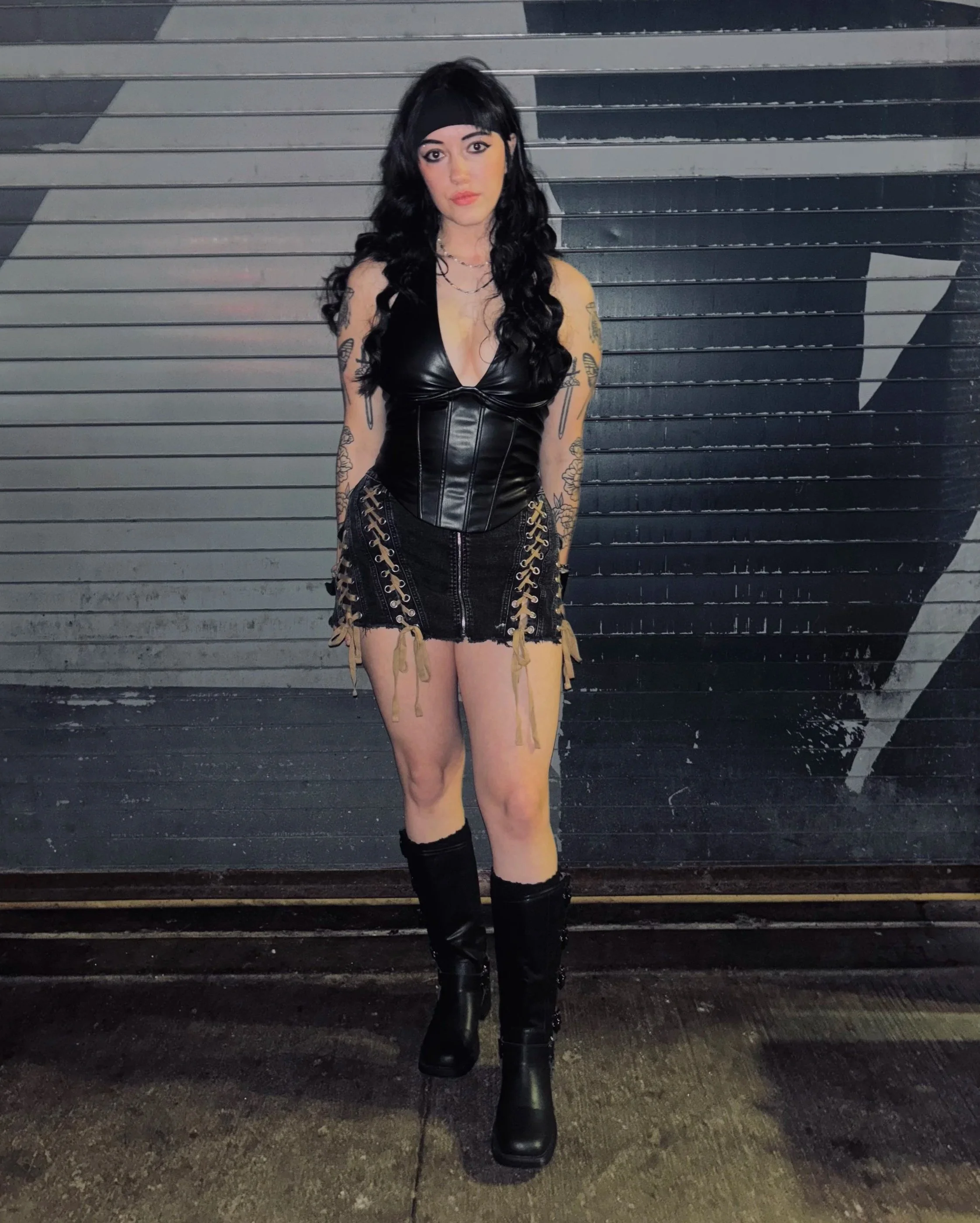 A woman with black, curly hair and tattoos on her arms, wearing a black headband, leather top, black shorts with lace-up sides, and black boots, standing in front of a gray and black metal garage door.