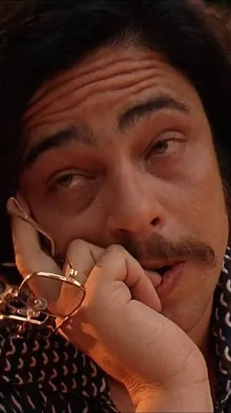 A man with dark hair and a mustache resting his chin on his hand, with a relaxed expression and slightly sleepy eyes