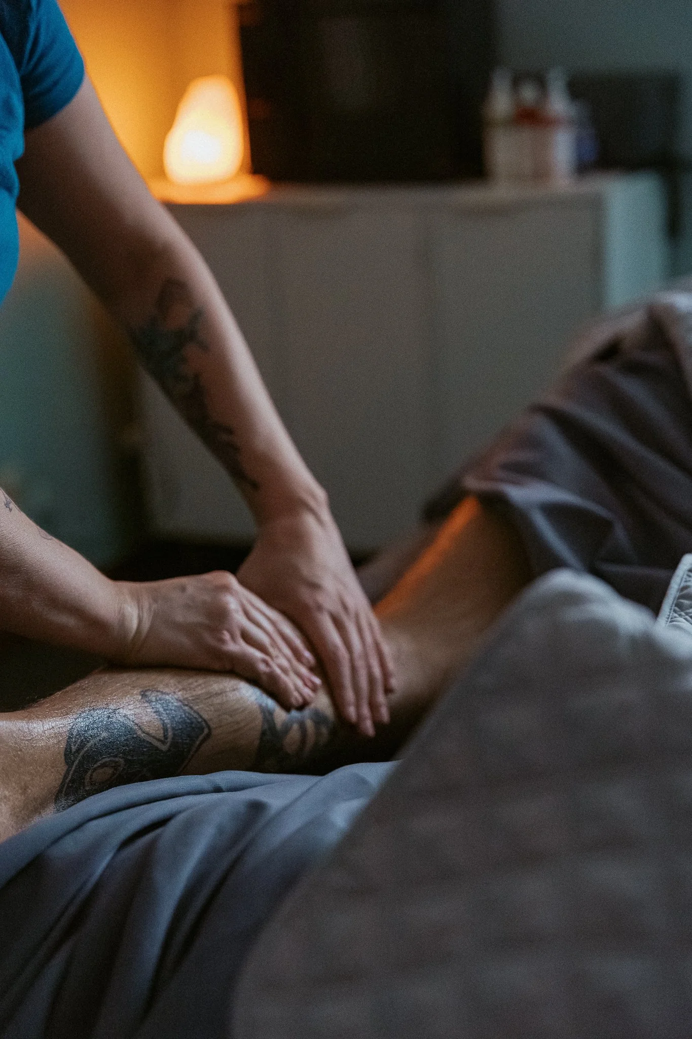 Close-up of a person receiving a massage on their leg from a massage therapist in a dimly lit room with a salt lamp on a nightstand in the background.