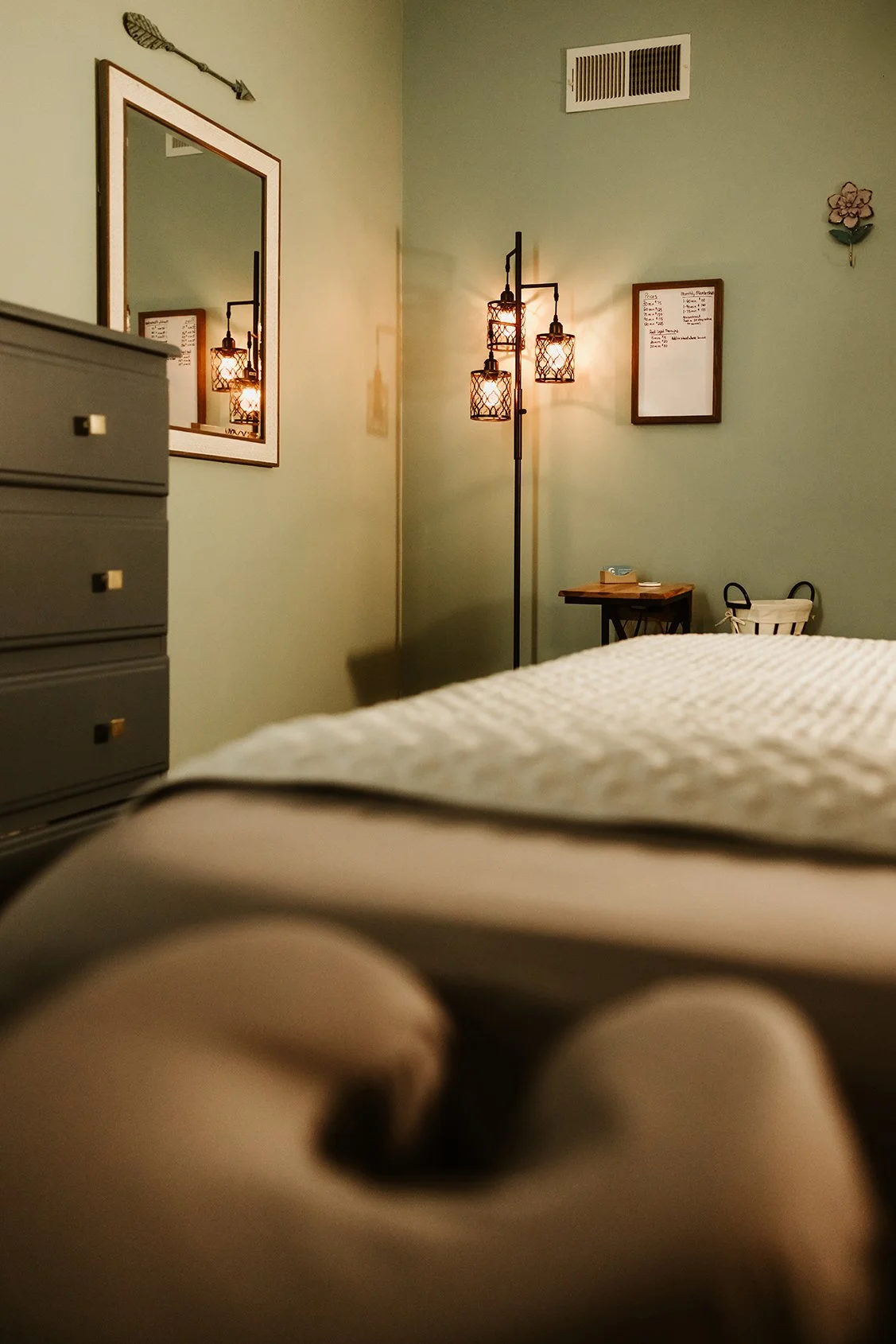 A bedroom with a bed in the foreground, a dresser on the left, a mirror on the wall, a lamp with three light fixtures, a small table with a tissue box, and a whiteboard on the wall.