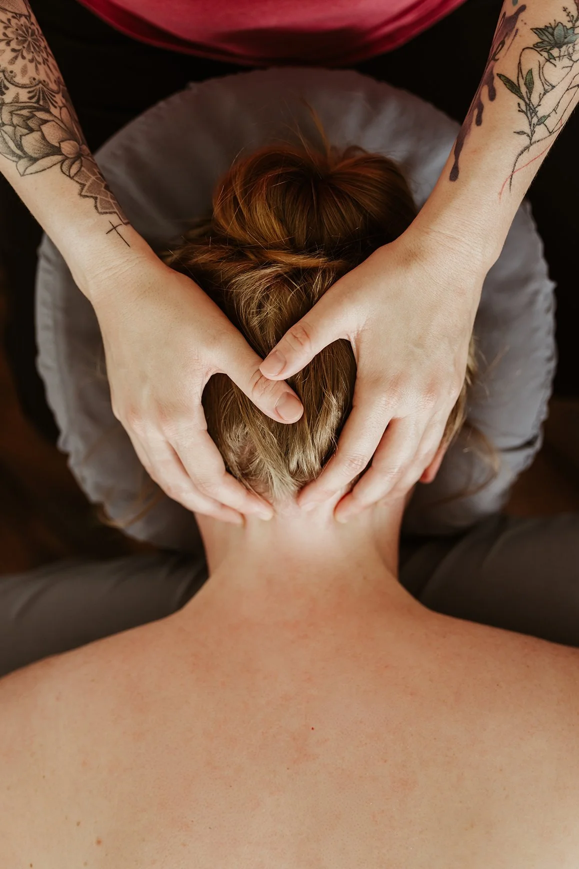 Person receiving a massage, lying face down with hands forming a heart shape behind their head, with tattoos visible on arms.