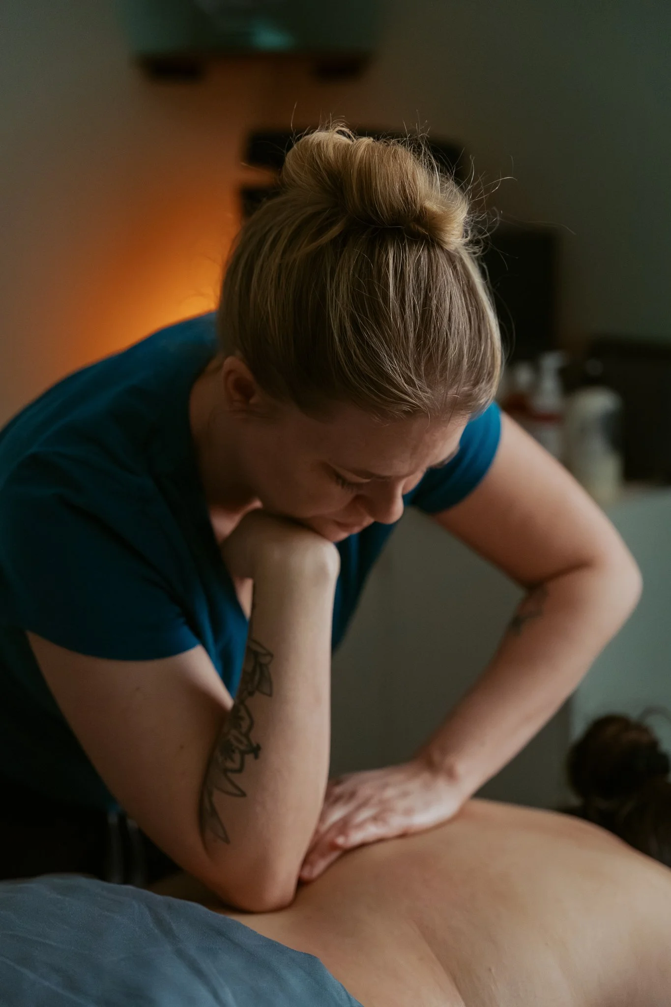 A woman with blonde hair styled in a bun, wearing a blue shirt, is leaning over and performing a massage or physical therapy on a person's leg in a dimly lit room.