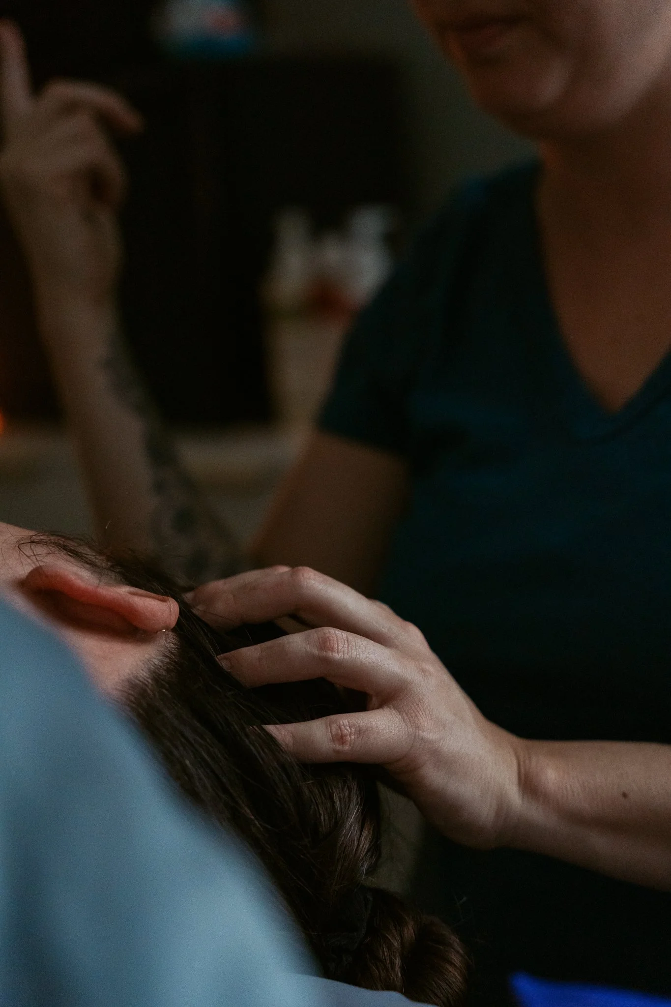 A person with long brown hair is resting their head on a hand, with another person leaning over and smiling nearby. The scene appears to be in a dimly lit setting.