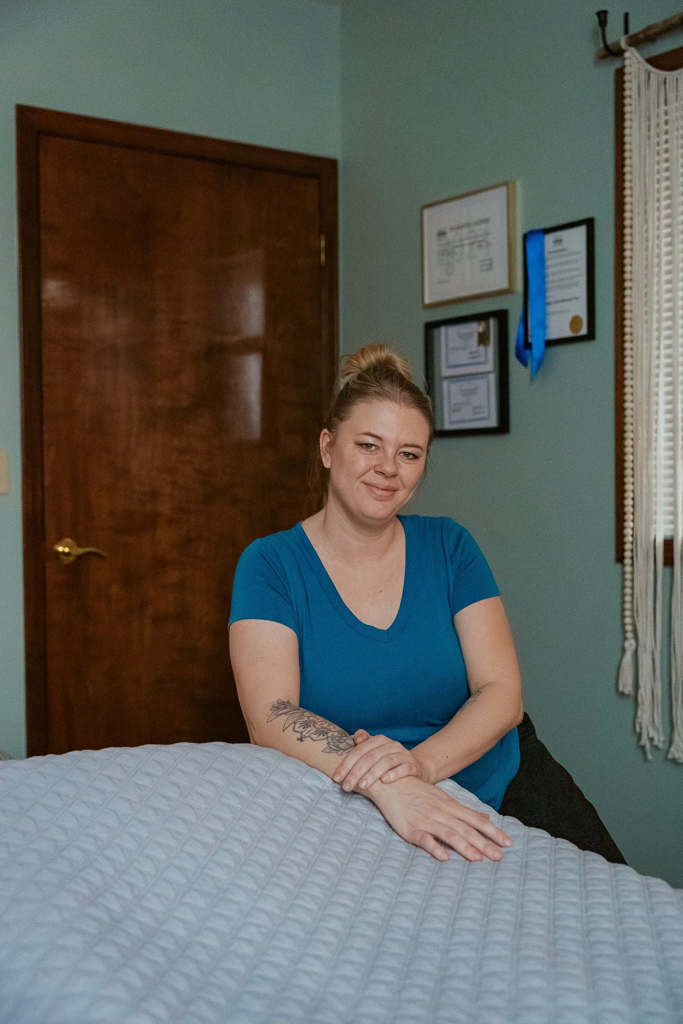 A woman in a blue shirt sitting at a bed with a white quilt. She has her arms crossed on the bed and a tattoo on her right forearm. The background shows a door, framed certificates on the wall, a window with lace curtains, and a green wall.