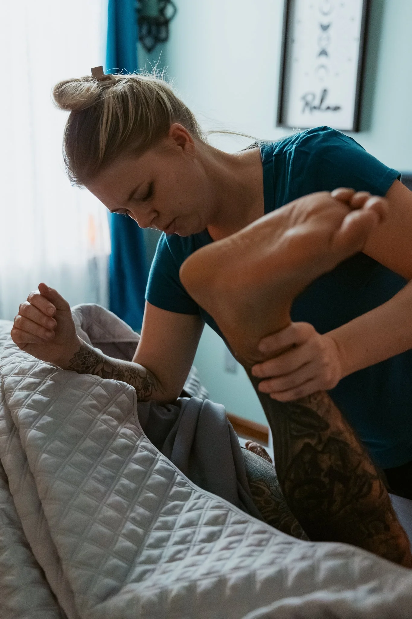 A woman doing a shoulder exercise with a tattooed arm holding her shoulder, while she is lying on a bed. A framed poster with the words 'Relax' and a decorative pattern are visible in the background.