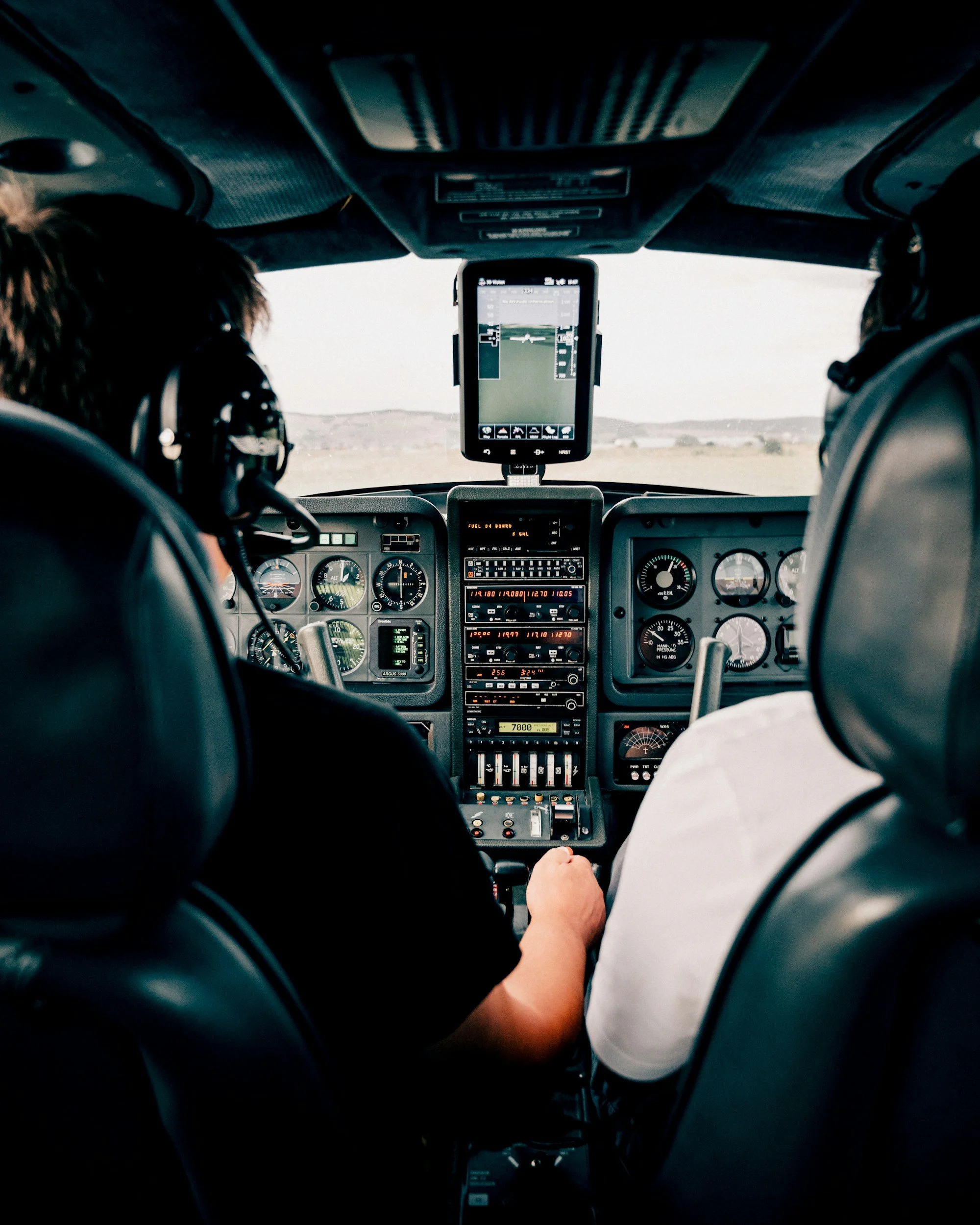 Interior view of an airplane cockpit with two pilots, one in a black shirt and the other in a white shirt, using flight instruments and screens, looking out at a vast open landscape or sky.