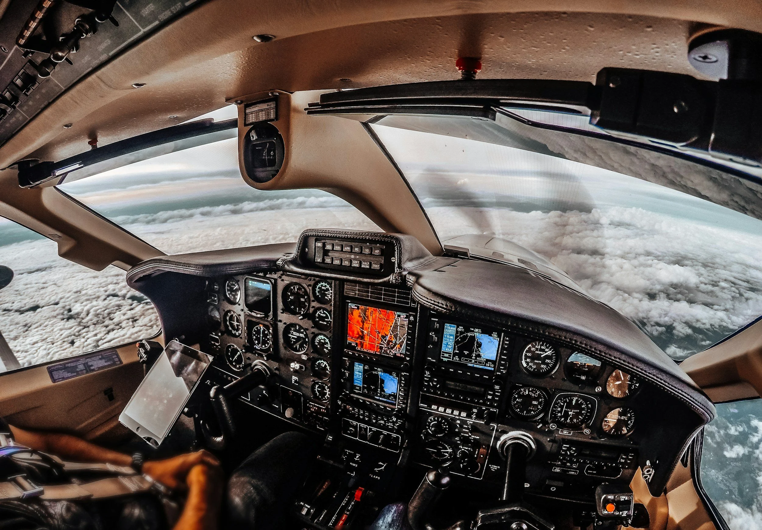 Cockpit view of an airplane flying above the clouds with various instrument panels, gauges, and navigation screens visible.