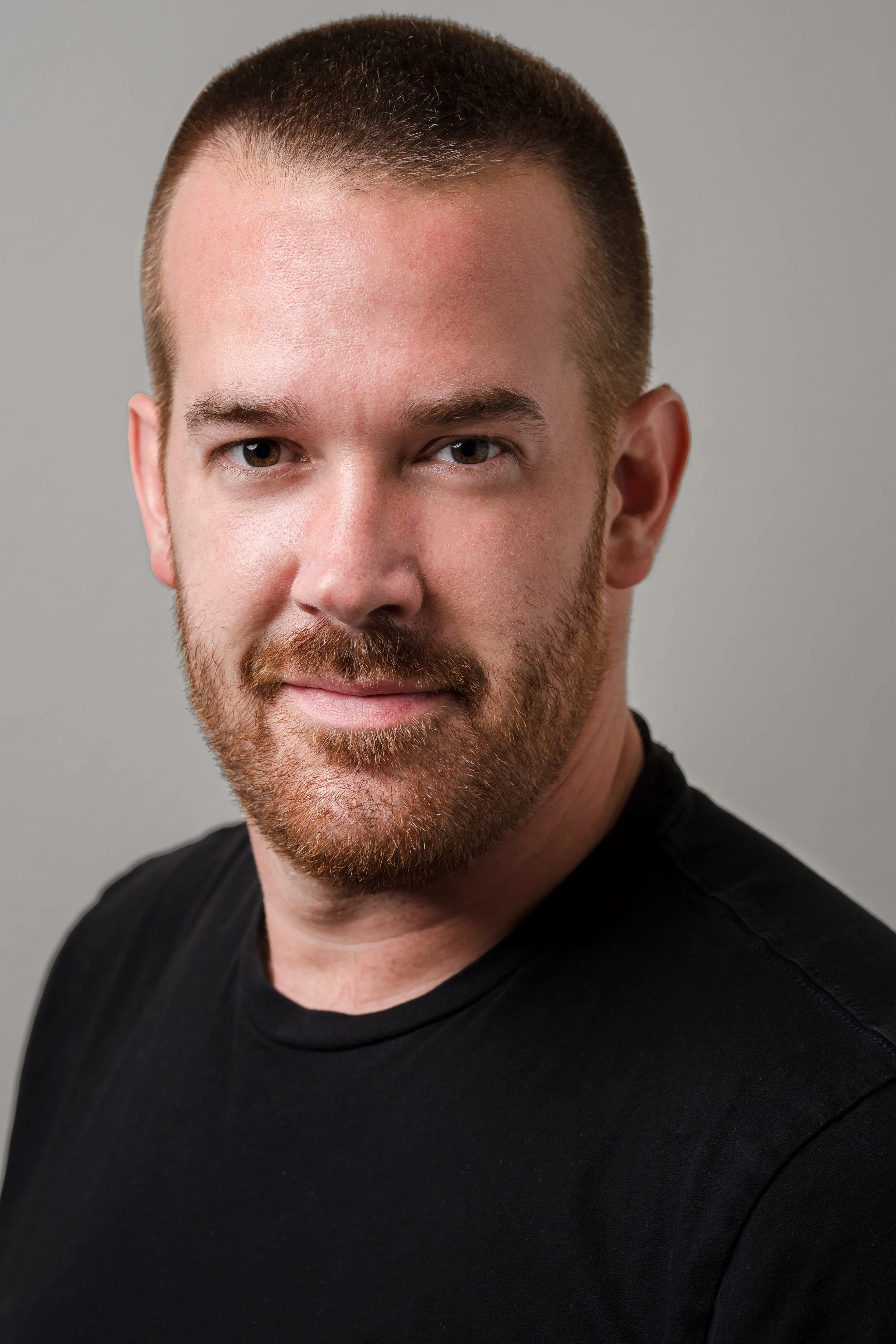 Close-up of a man with short brown hair, a light beard, wearing a black shirt, smiling slightly against a gray background.