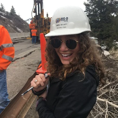 A woman smiling while at a construction site, wearing a white hard hat, sunglasses, and a black jacket, with construction workers and equipment in the background.