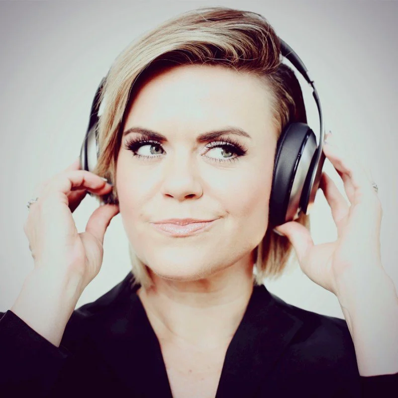 A woman with short blonde hair wearing headphones, holding them with both hands, and looking to the side with a confident expression.