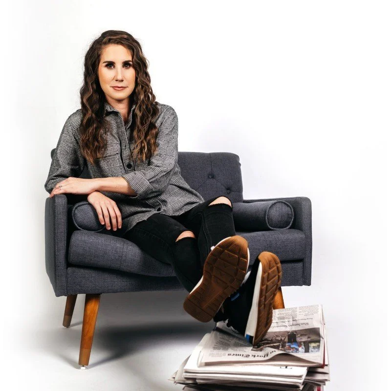 Young woman sitting on a gray sofa with her feet resting on a stack of newspapers, wearing a checkered shirt and ripped black jeans.