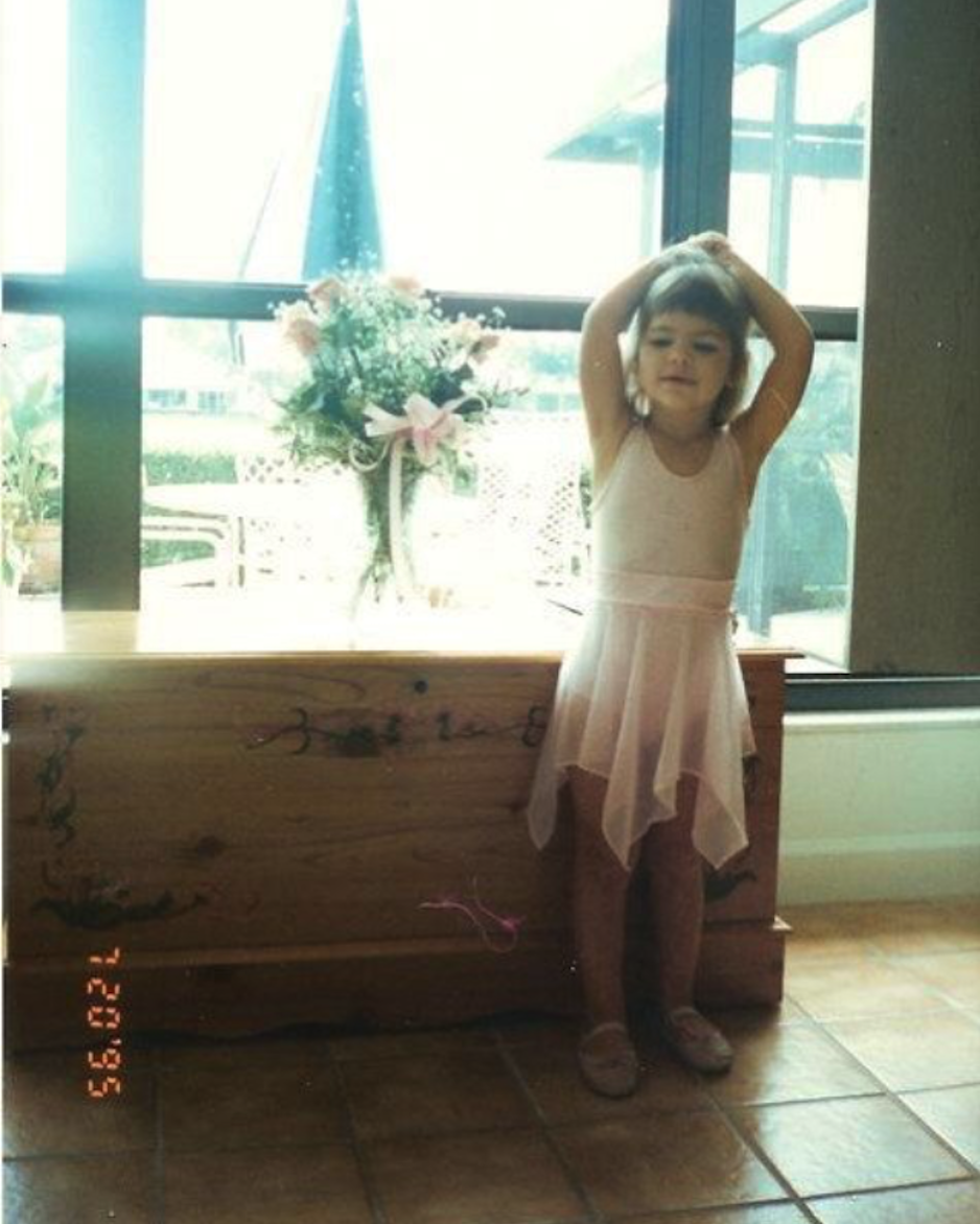 A young girl wearing a pink dress with her hands on her head standing next to a window with flowers and an outdoor patio in the background.