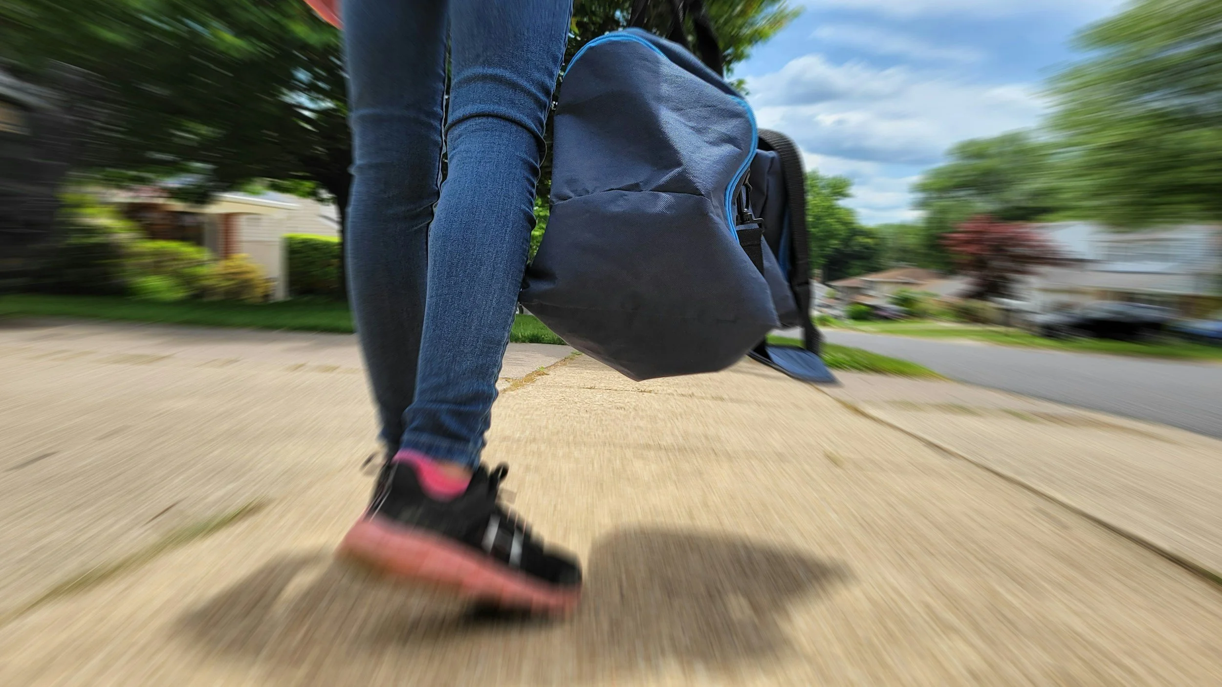 A person walking or running on a sidewalk carrying a blue backpack, with motion blur indicating movement, in a residential neighborhood with trees and houses under a partly cloudy sky.