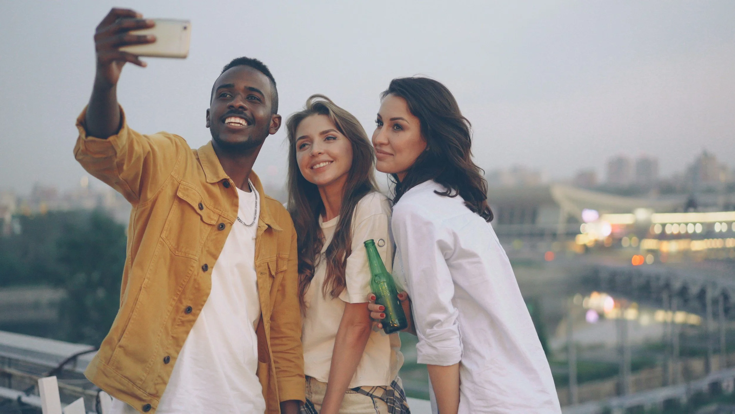 Three friends, two women and one man, taking a selfie together outdoors in the evening with a cityscape in the background. The man is holding a smartphone, and one woman is holding a green glass bottle.