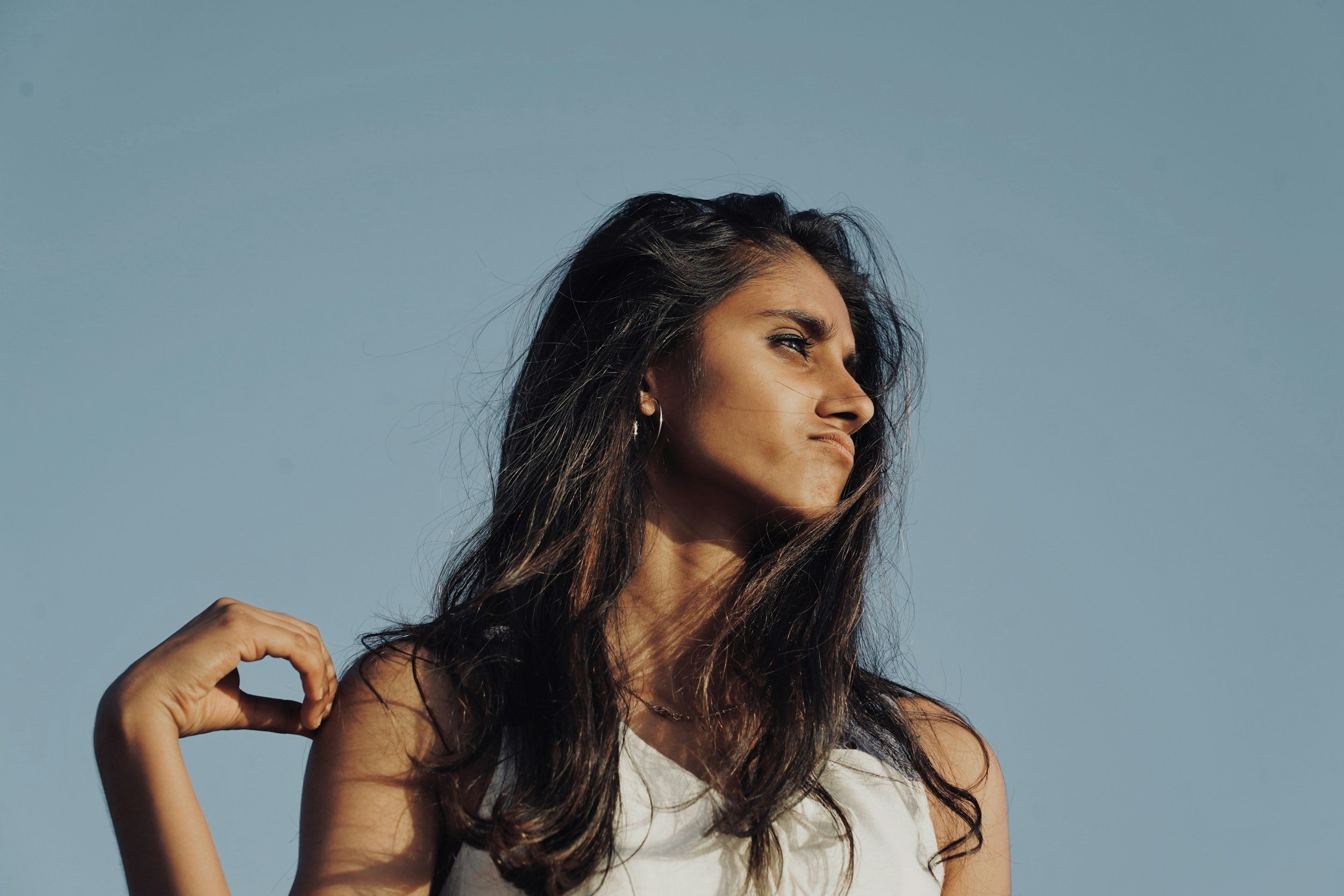 A woman with long brown hair outdoors against a clear blue sky, making a fist with her right hand and raising her arm, looking to the side.