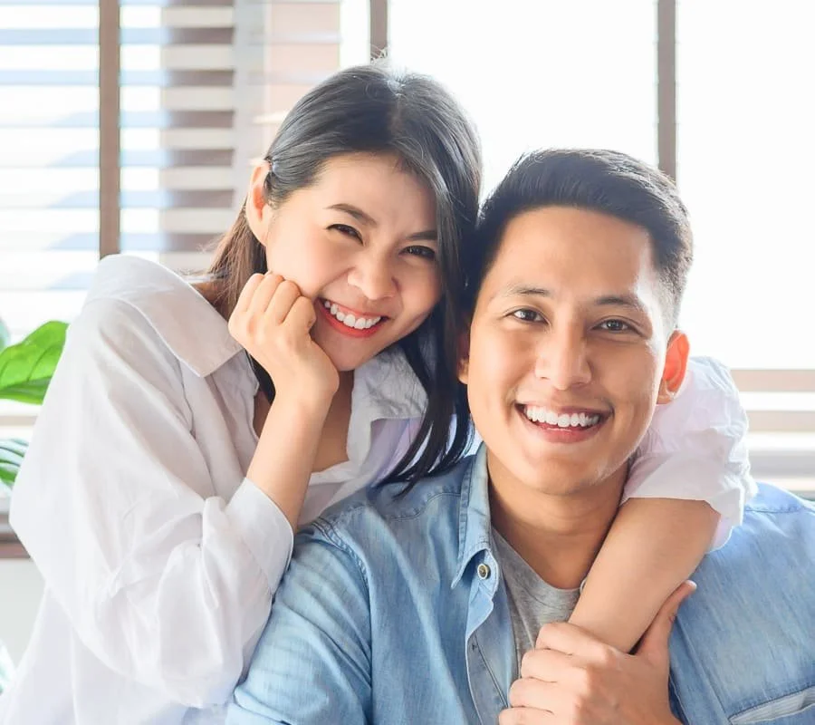 A smiling woman and man sit close together, with the woman leaning on the man's shoulder, indoors with sunlight coming through window blinds.