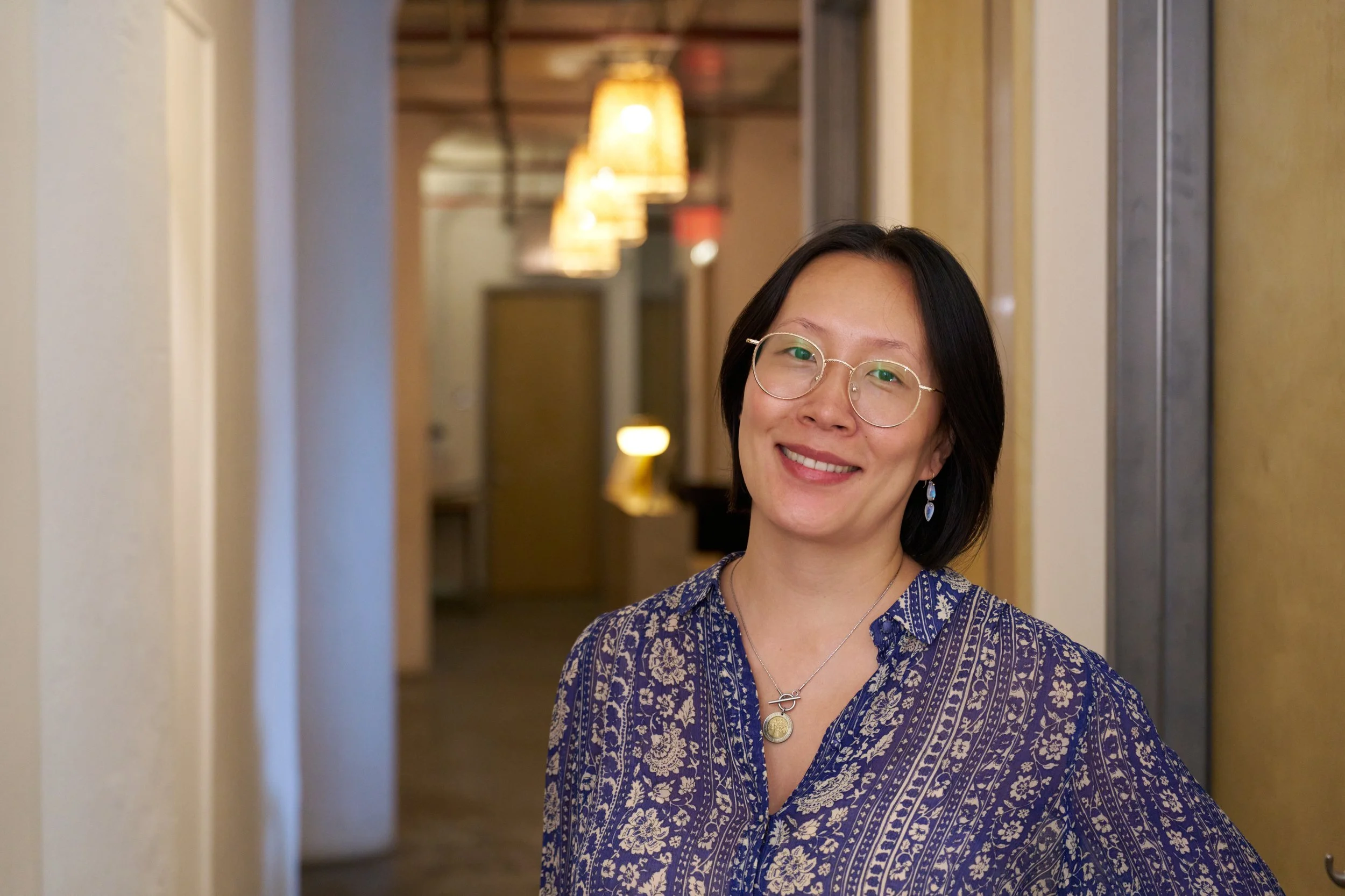 A woman with short dark hair and glasses smiling, wearing a blue patterned blouse and earrings, standing in an indoor hallway with warm lighting before couples therapy in NYC.