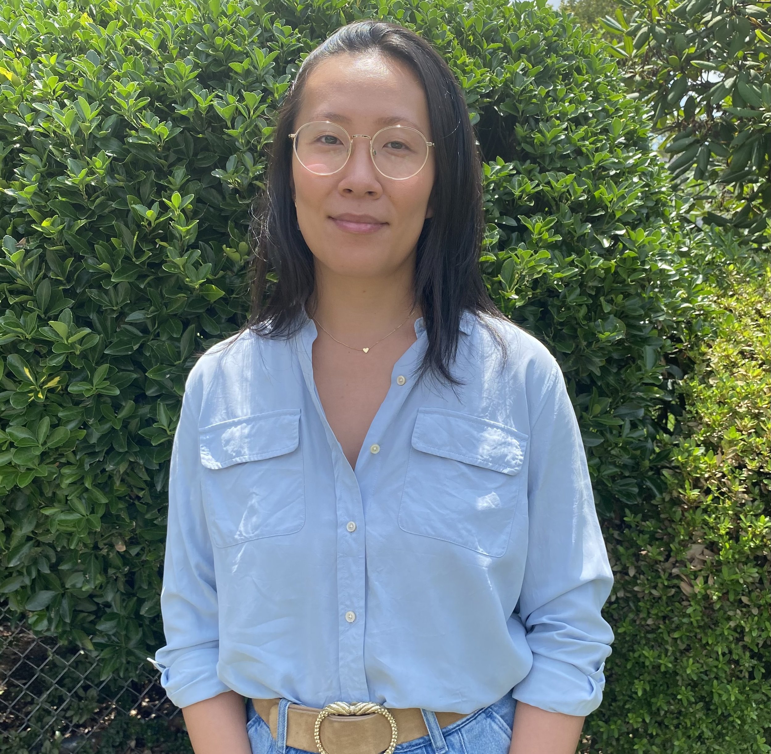 A woman with long black hair wearing glasses and a light blue shirt, standing in front of green bushes, smiling gently.