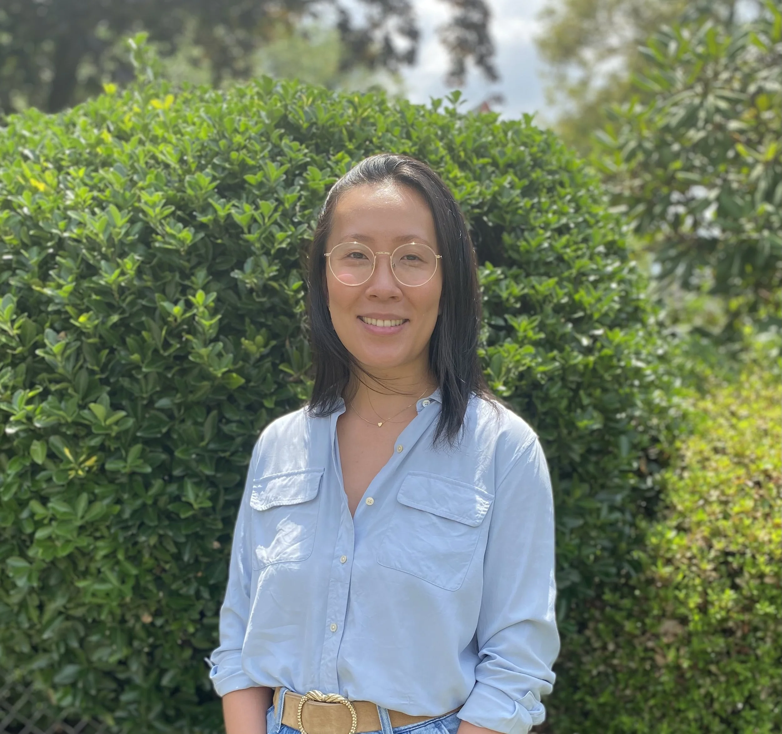 A woman with glasses smiling outdoors, standing in front of green bushes with trees in the background, wearing a light blue shirt and a gold bracelet.