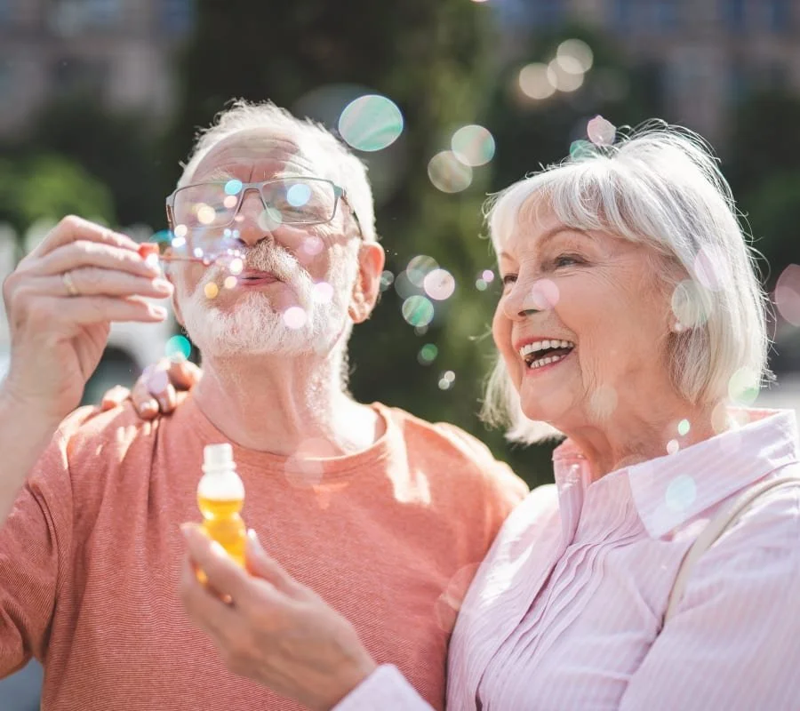 Smiling elderly couple outdoors with bubbles, man holding bubble solution and blowing bubbles, woman laughing.