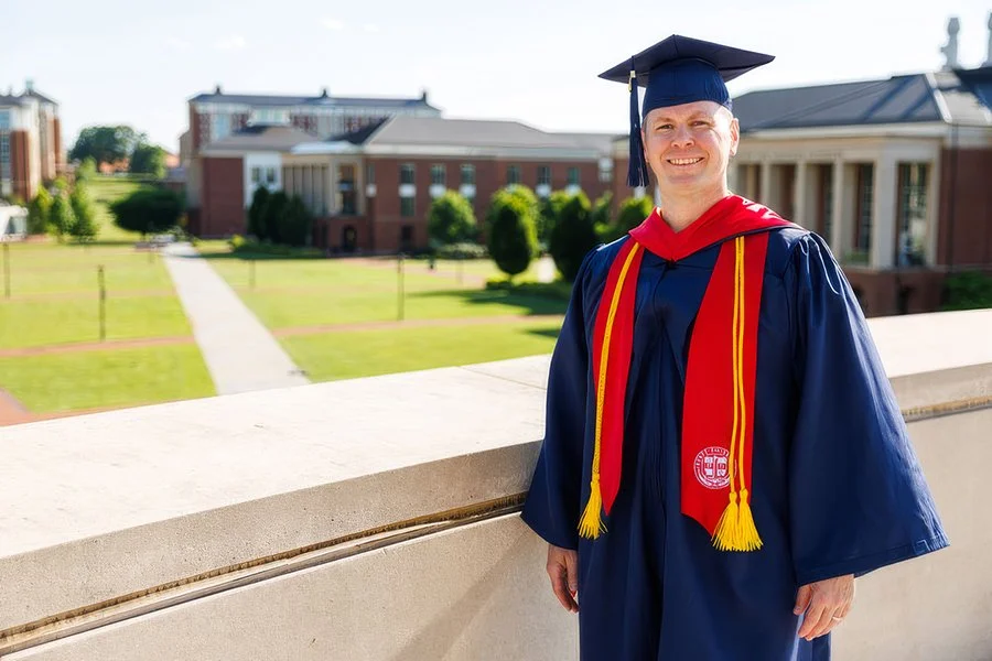 A smiling graduate in a navy cap and gown with a red and yellow stole, standing outdoors on a college campus with brick buildings and green lawns.