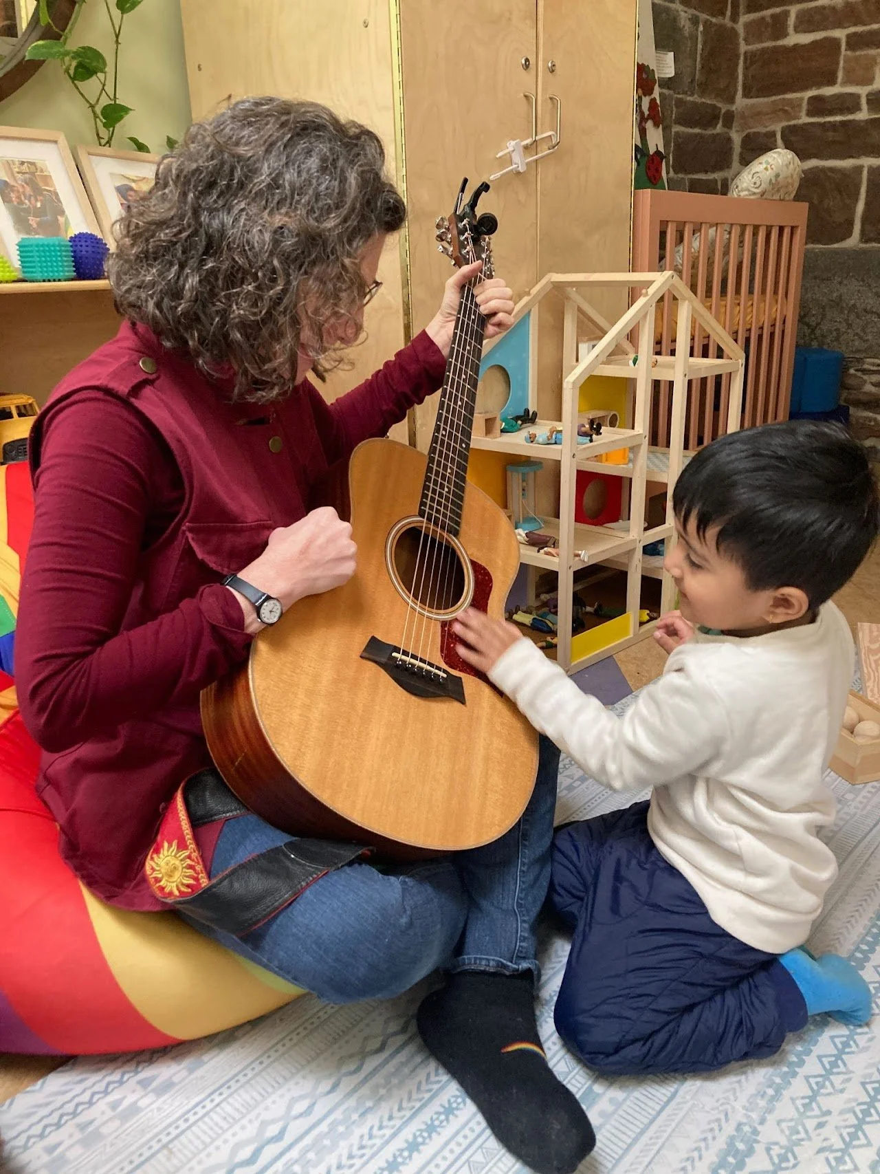 A woman with curly gray hair playing an acoustic guitar while a young boy with black hair touches the guitar strings, in a playroom with shelves of toys and framed pictures.