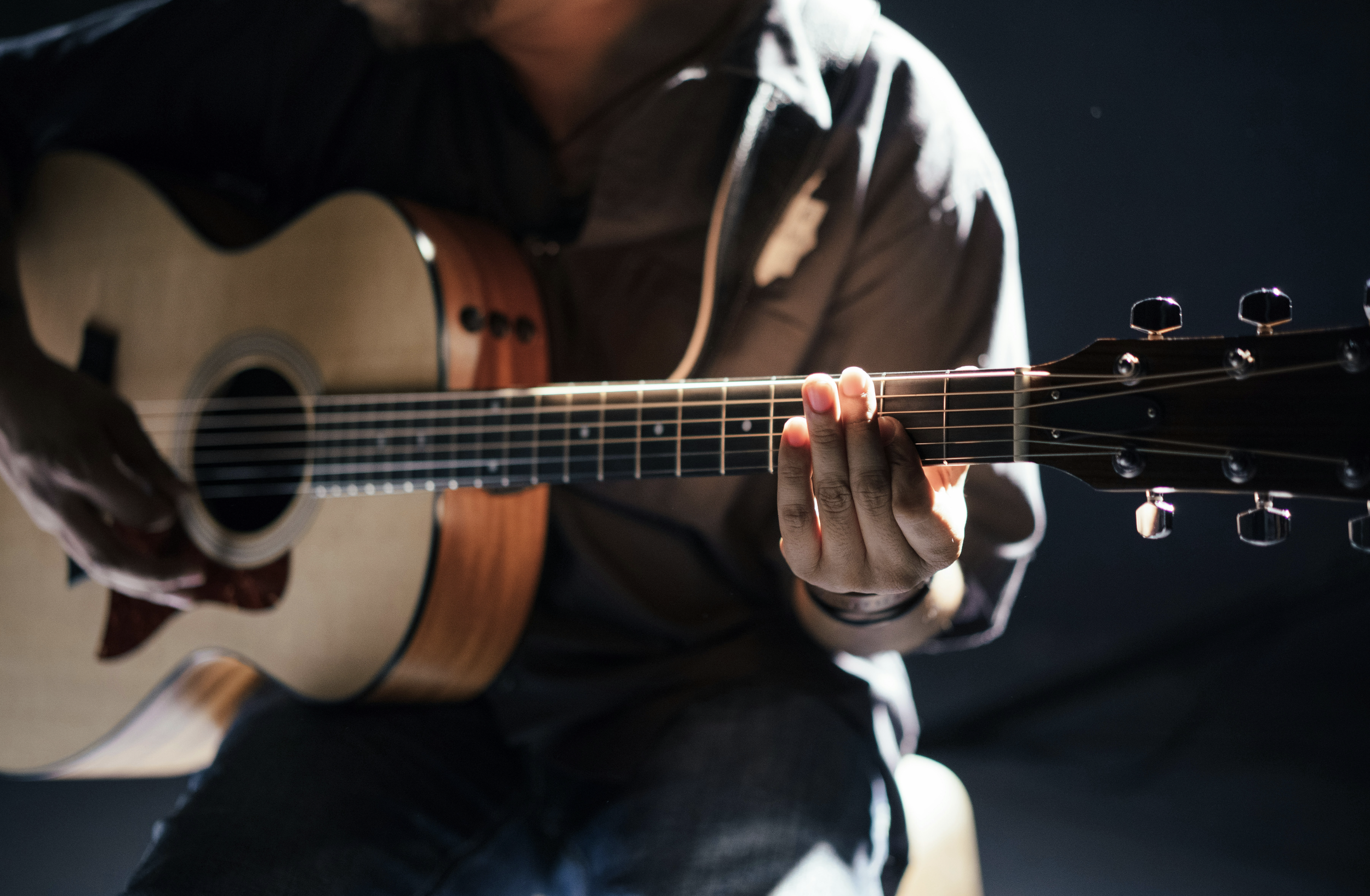 Person playing an acoustic guitar, focusing on their hand on the fretboard and part of the guitar's body, wearing a dark jacket with a shirt underneath, in a dimly lit setting.