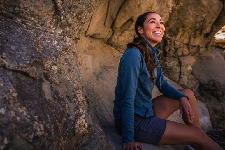 Victoria Gracie smiling while sitting among natural rock formations outdoors, wearing a blue jacket and shorts.