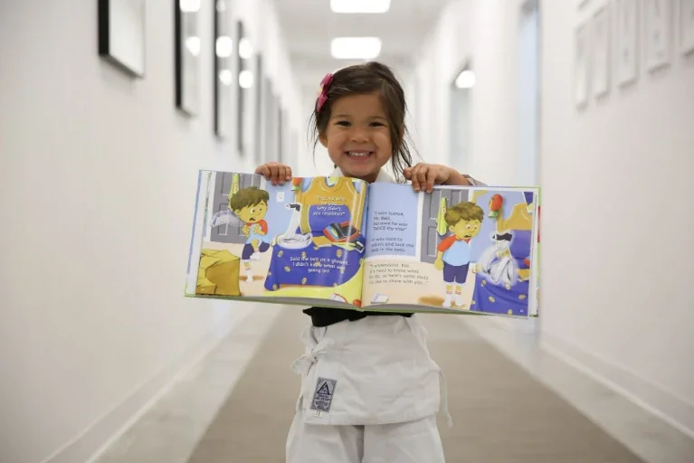 Young girl wearing a white Brazilian jiu jitsu uniform smiling while holding an open illustrated children's book in a bright hallway.