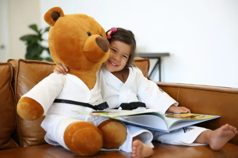 Young girl in a white Brazilian jiu jitsu uniform smiling beside a teddy bear wearing a matching uniform while reading a picture book on a brown leather couch.