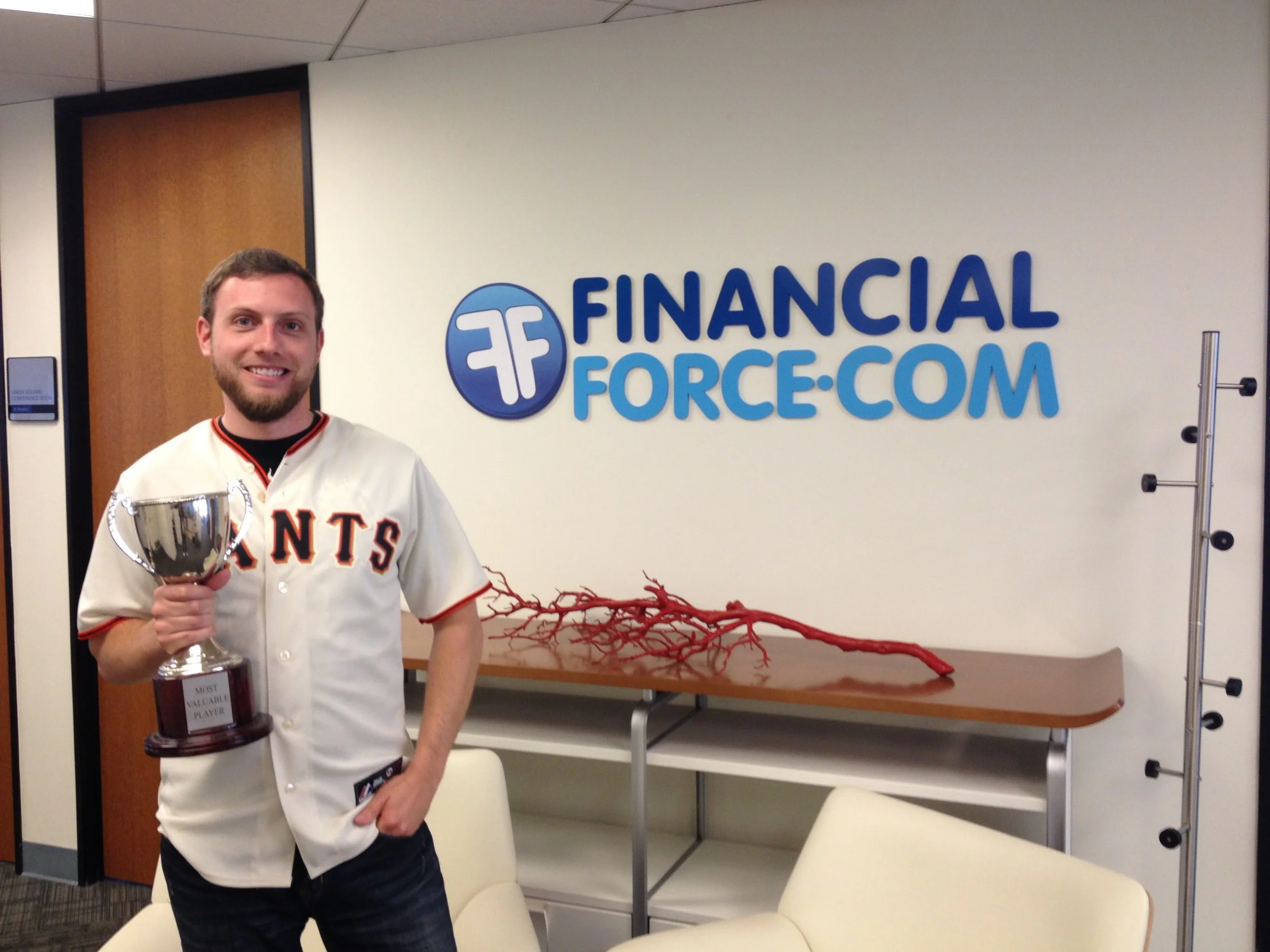 A man with a beard and short hair standing in front of a sign that reads 'FinancialForce.com.' He is smiling and holding a trophy that says 'Most Valuable Player.' There are red branches or coral on a table behind him, in an office setting.
