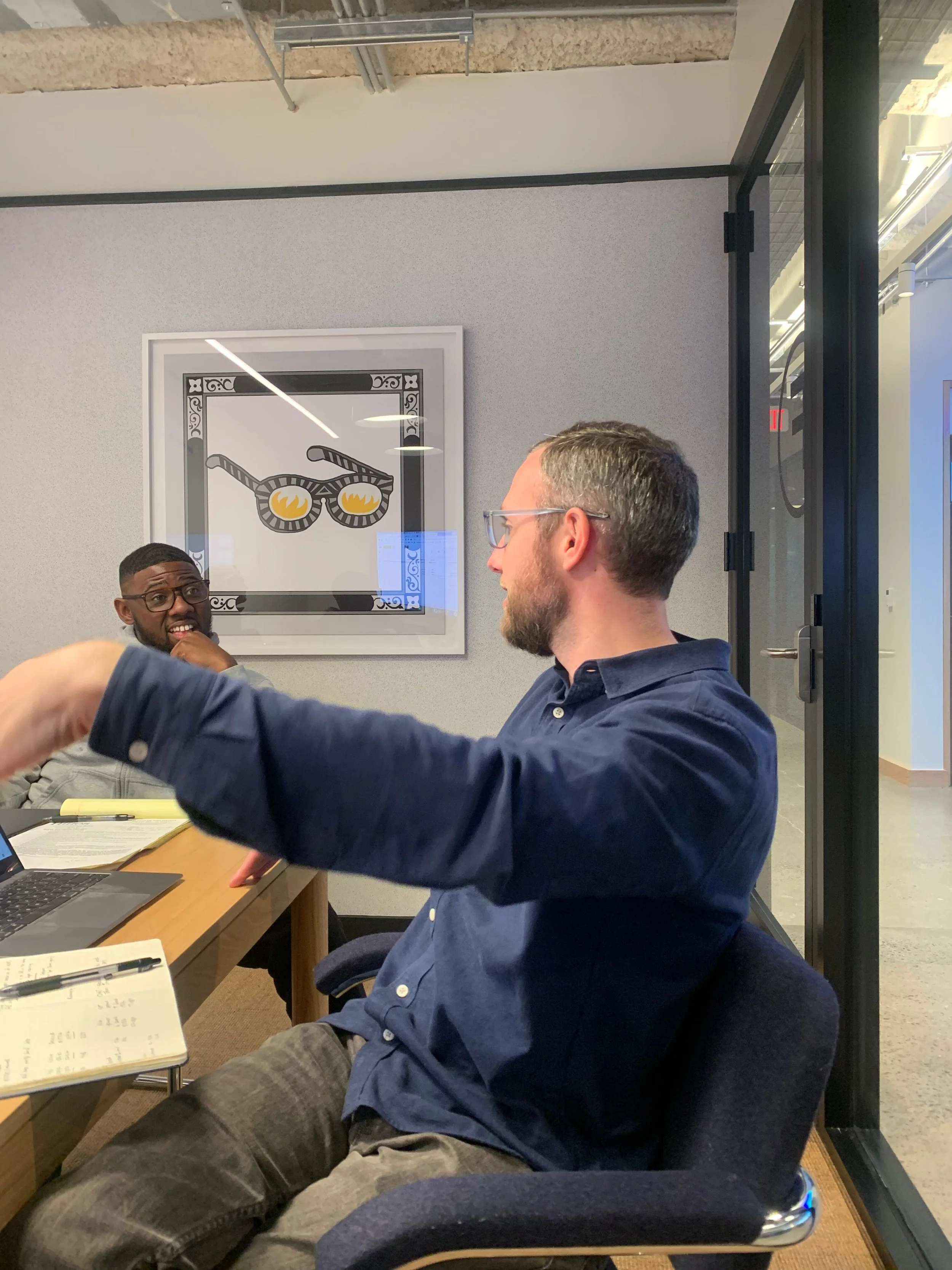 Two men engaged in conversation at a table in a modern office or conference room. One man, with glasses and a beard, is sitting in a rolling chair, while the other man, with a short haircut and glasses, appears to be speaking or responding. On the wall behind them is framed art of sunglasses with flames in the lenses.