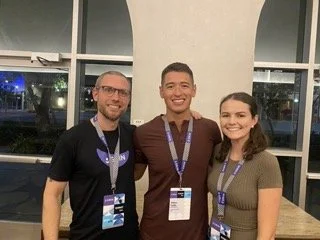 Three people smiling and standing together at an event, wearing conference badges, inside a modern building with large windows.