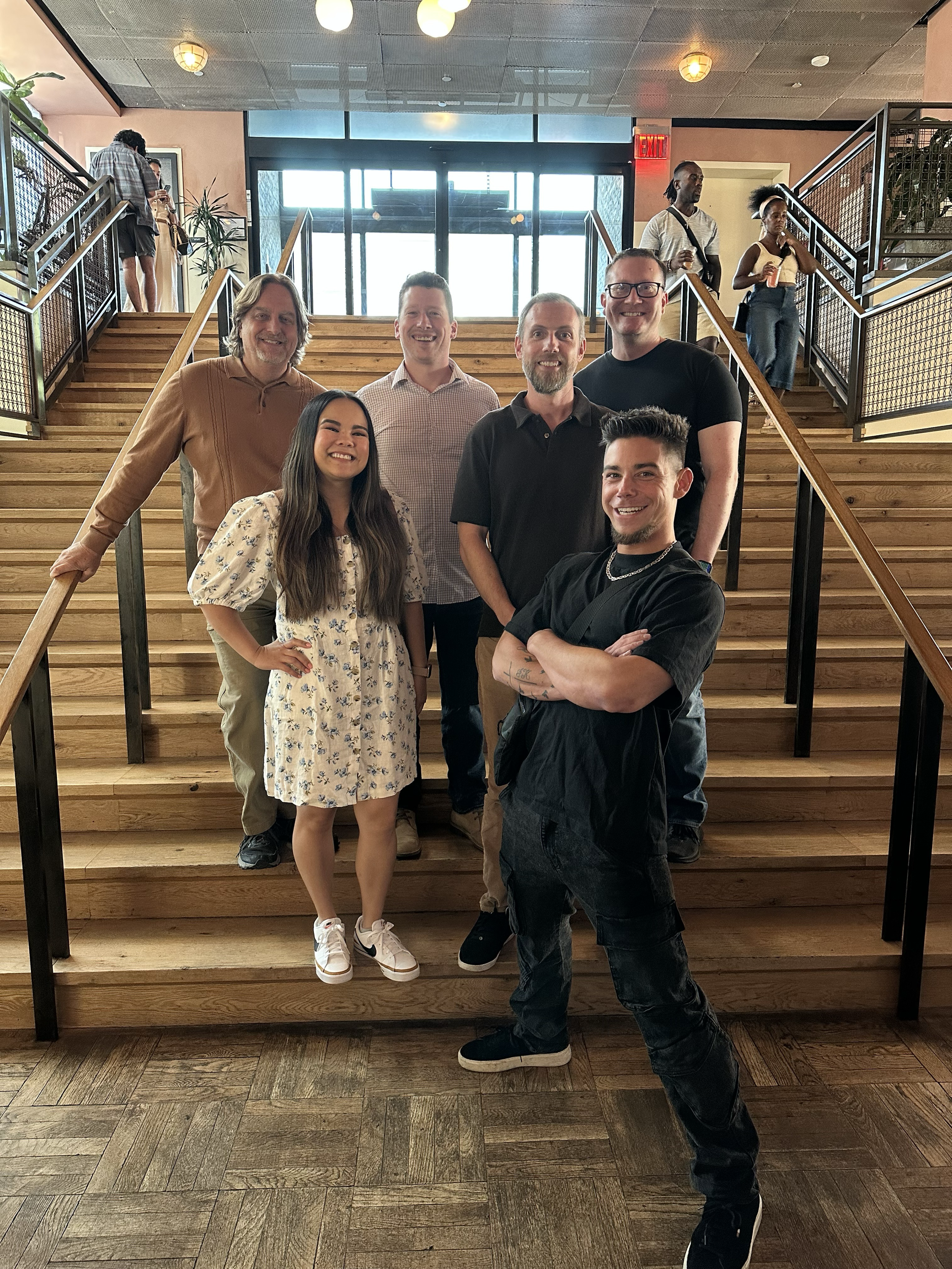 A group of six people posing on a wooden staircase inside a building with large glass windows. Four people are in the front row, smiling at the camera, while two are on the stairs in the background, one taking a photo and the other holding a drink. The staircase has black metal railings and the ceiling has hanging lights.